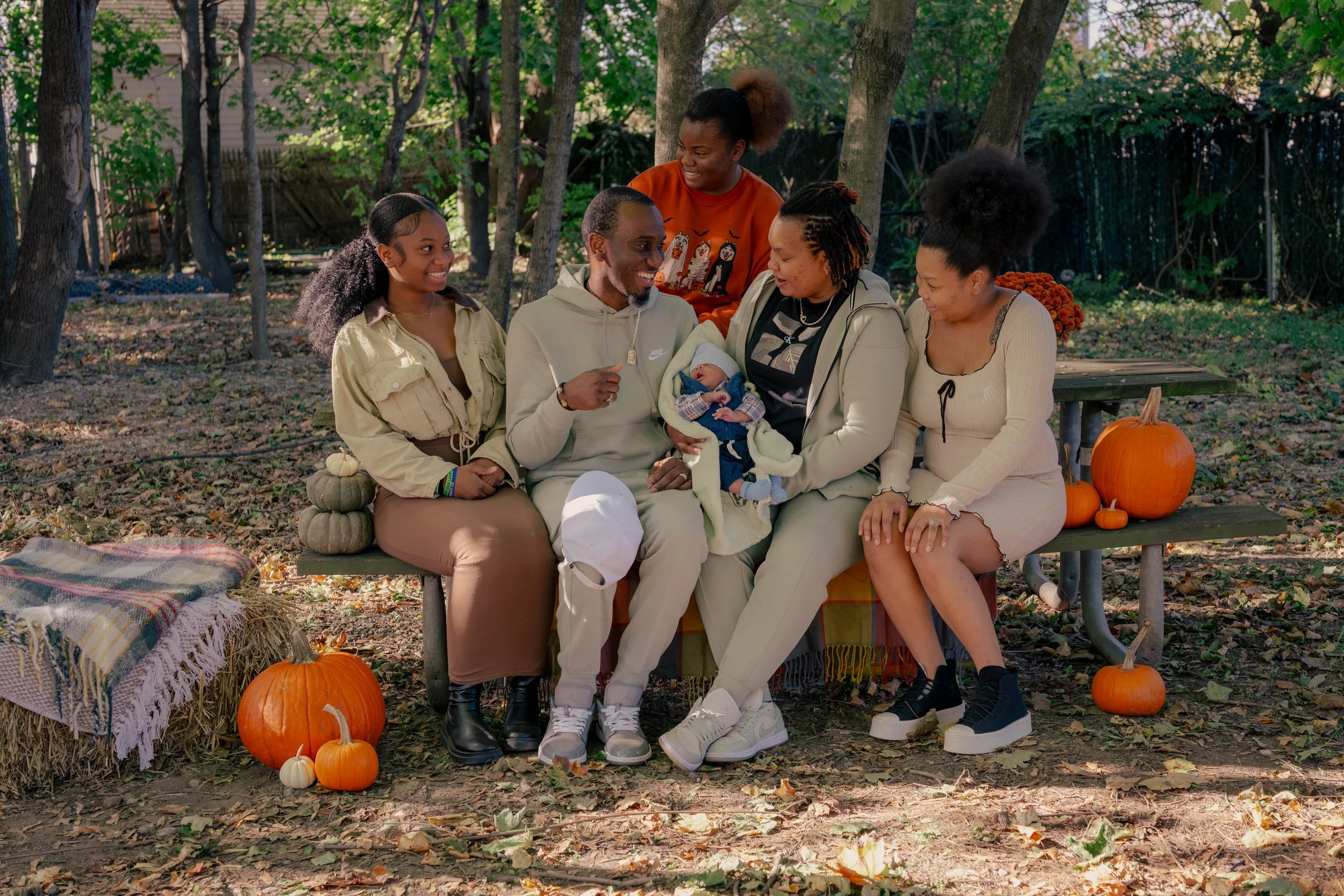A multiracial group of six people, including a baby, sitting on a bench outdoors surrounded by autumn leaves and pumpkins, enjoying a fall gathering.