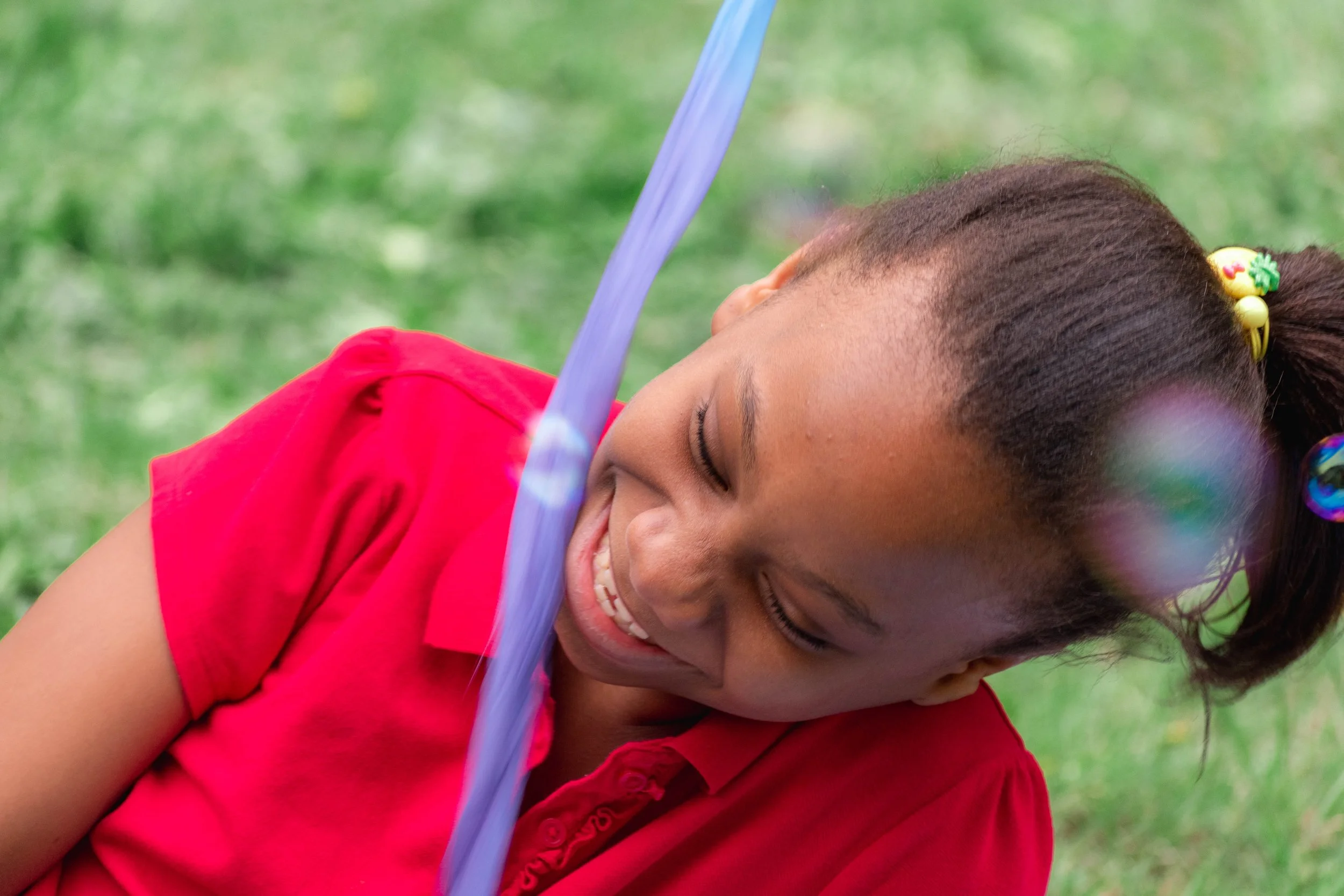 Young girl laughing and playing outdoors on green grass, wearing a red shirt and colorful hair ties.