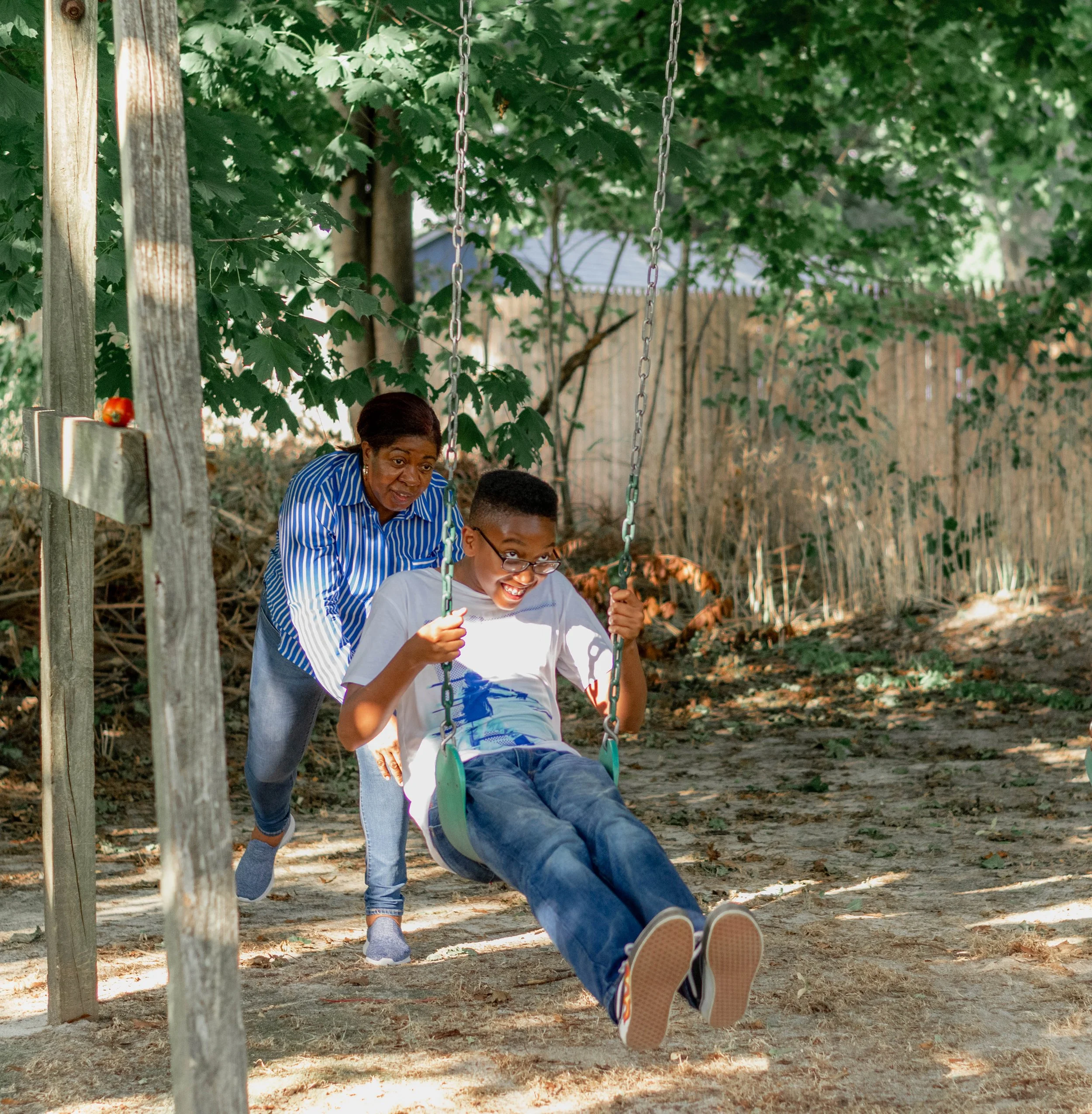 An elderly woman pushing a young boy on a swing in a backyard with trees and a wooden fence.