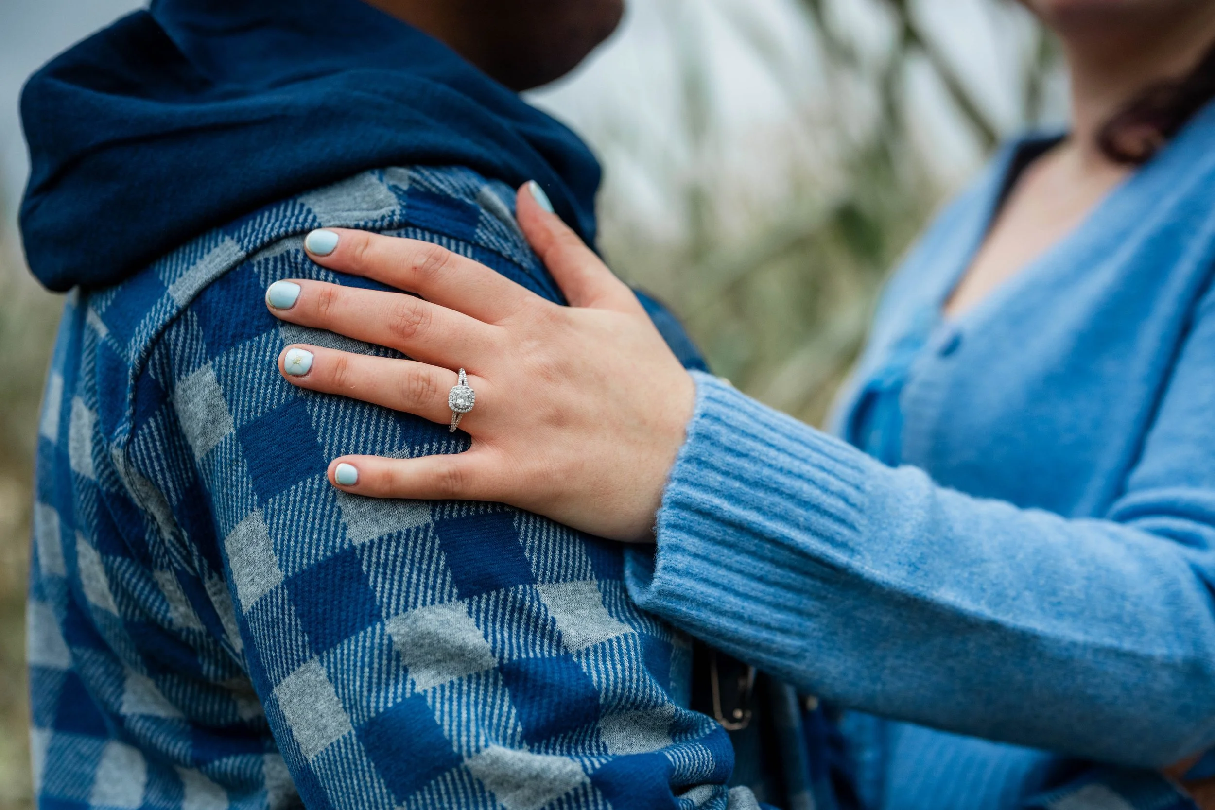 Close-up of a woman placing her hand on a man's shoulder, showing an engagement ring with a large central diamond surrounded by smaller diamonds, against a background of outdoors with blurred greenery.