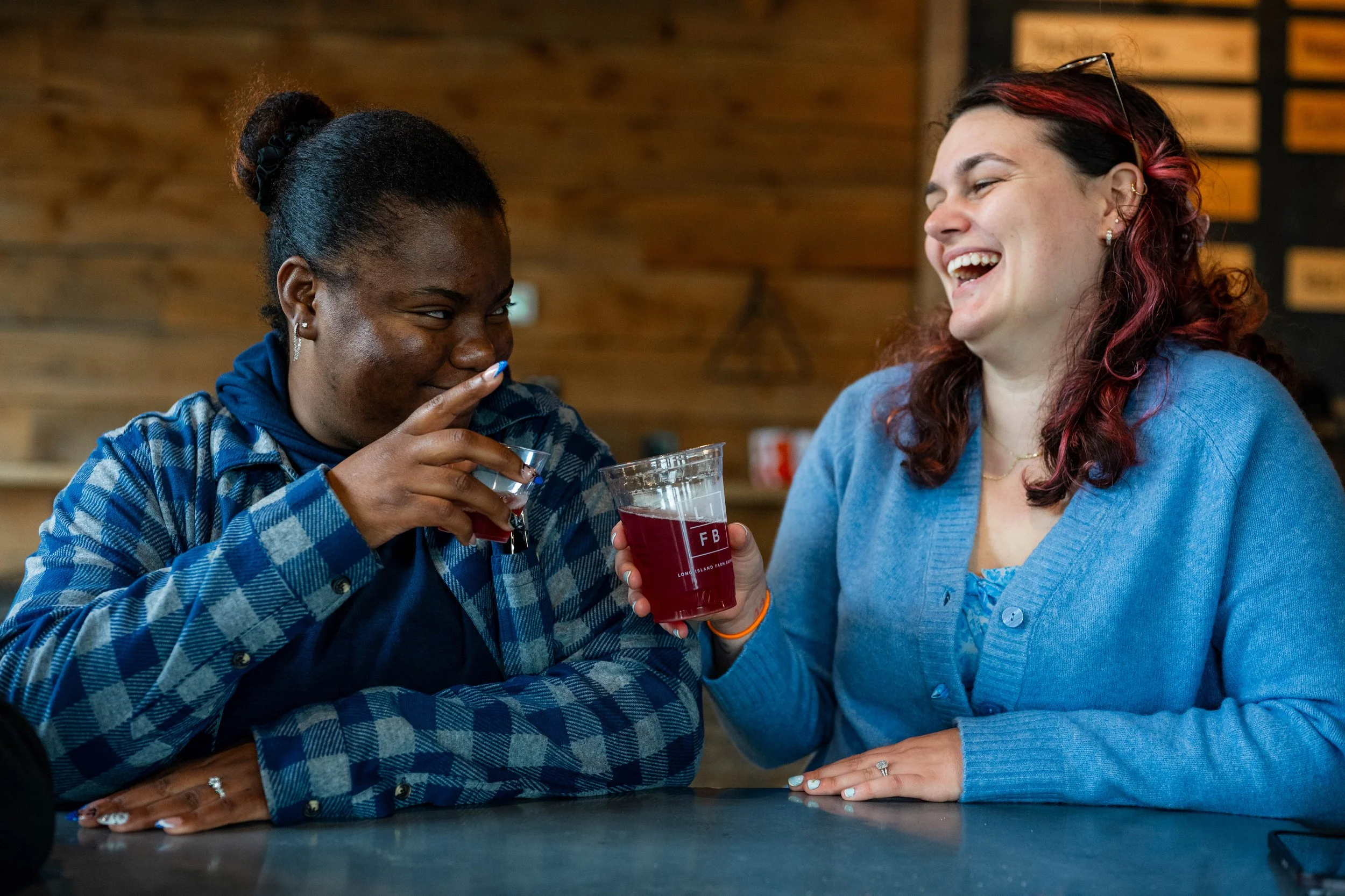 Two women amusement and laughter, holding glasses with dark red drinks, sitting at a bar or cafe with wooden wall background.