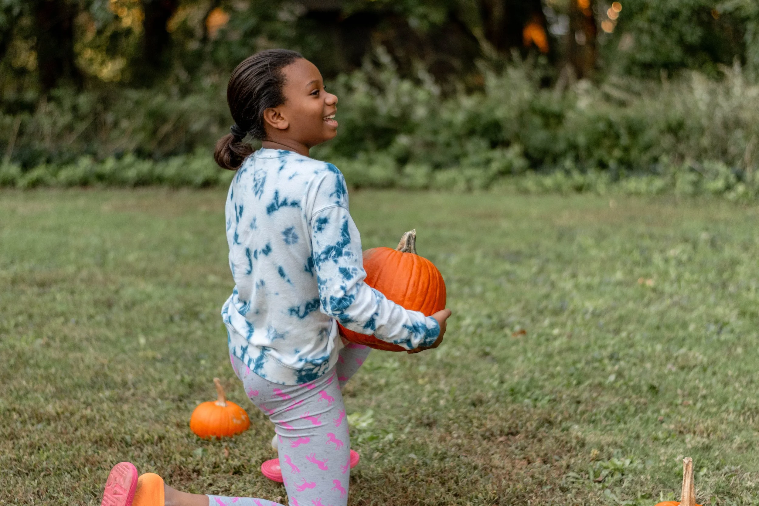 A young girl with braided hair, wearing a tie-dye sweatshirt and pink leggings, is outdoors in a grassy area holding a large orange pumpkin. There are smaller pumpkins on the ground nearby, and she is smiling while looking to the side.