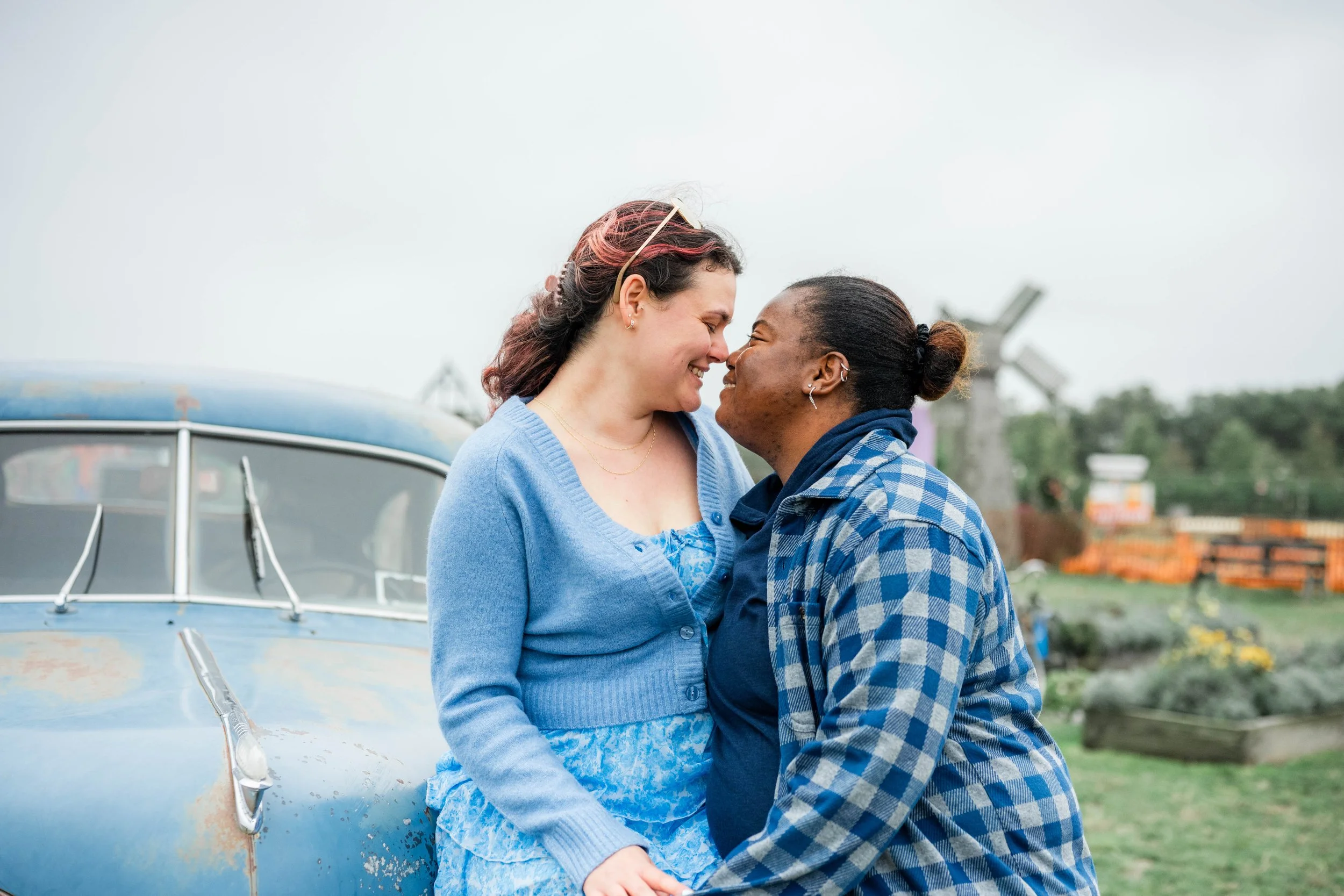 Two women face each other with noses touching, smiling happily, outdoors near a vintage blue car, with a garden and a windmill in the background.