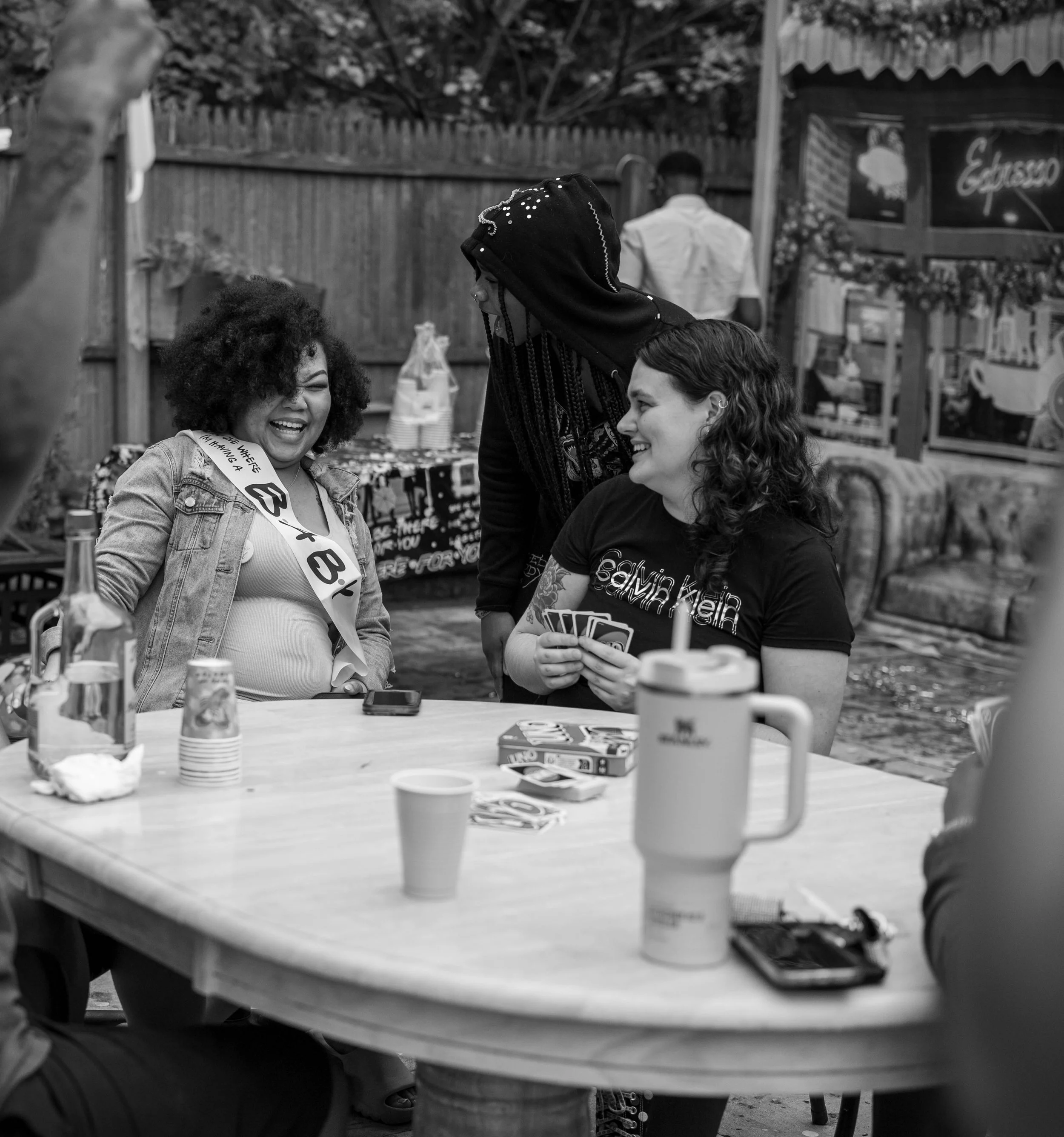 Three women at a table outdoors, laughing and playing cards, with drinks and snacks on the table.