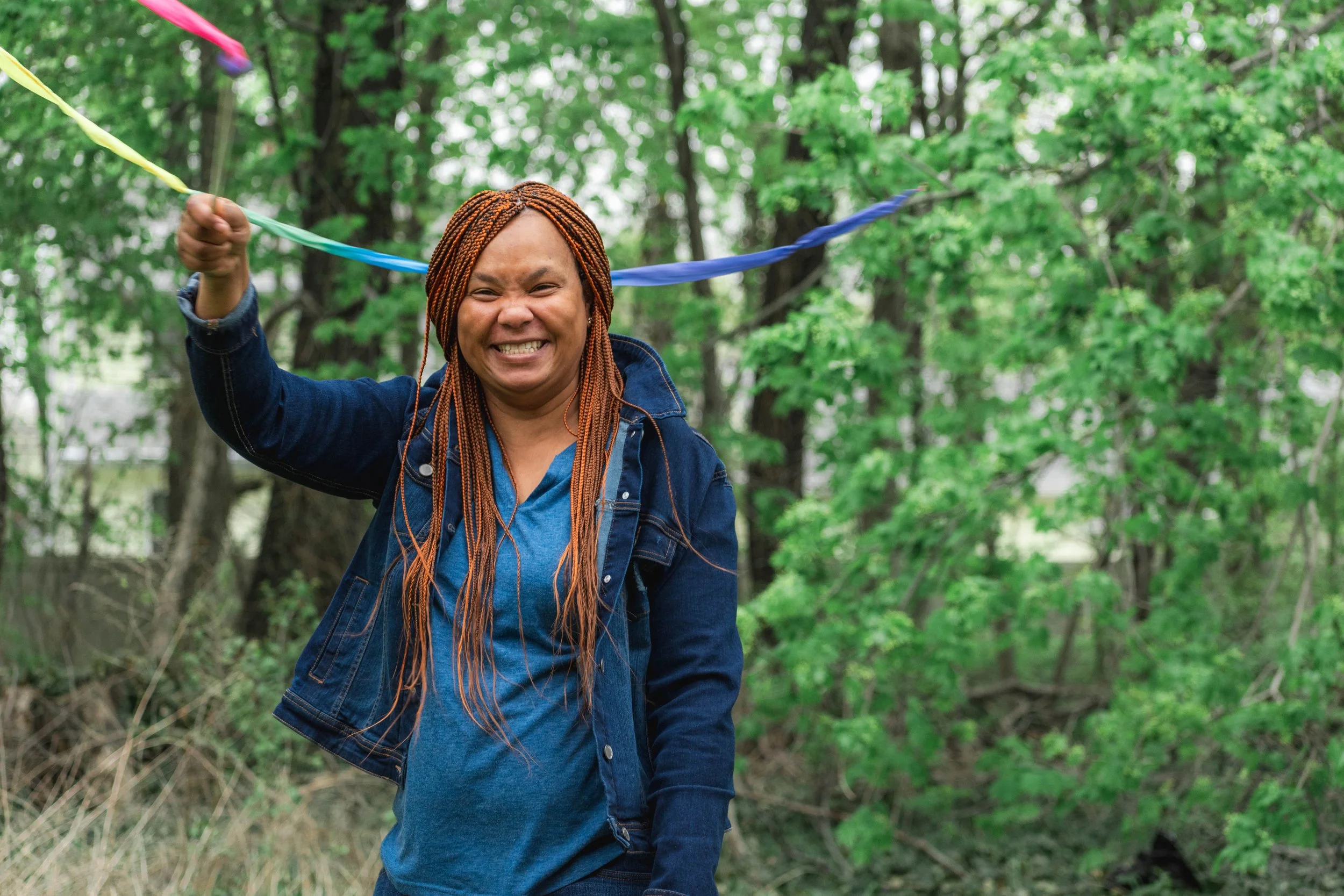 A smiling woman with braided hair holding a string of colorful flags outdoors in a green, wooded area.