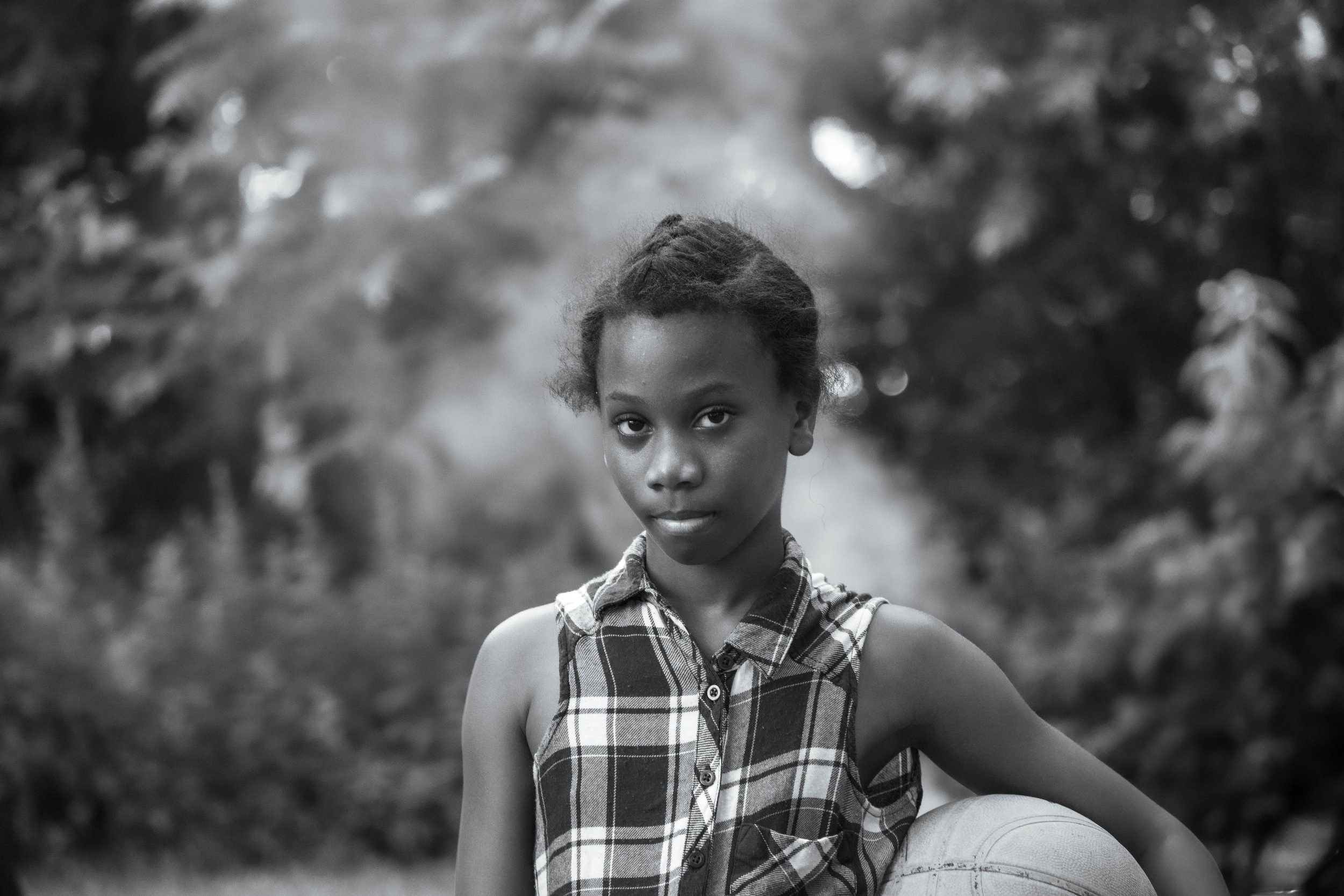 A young girl with short natural hair wearing a sleeveless plaid shirt, holding a basketball, outdoors with blurred trees in the background.
