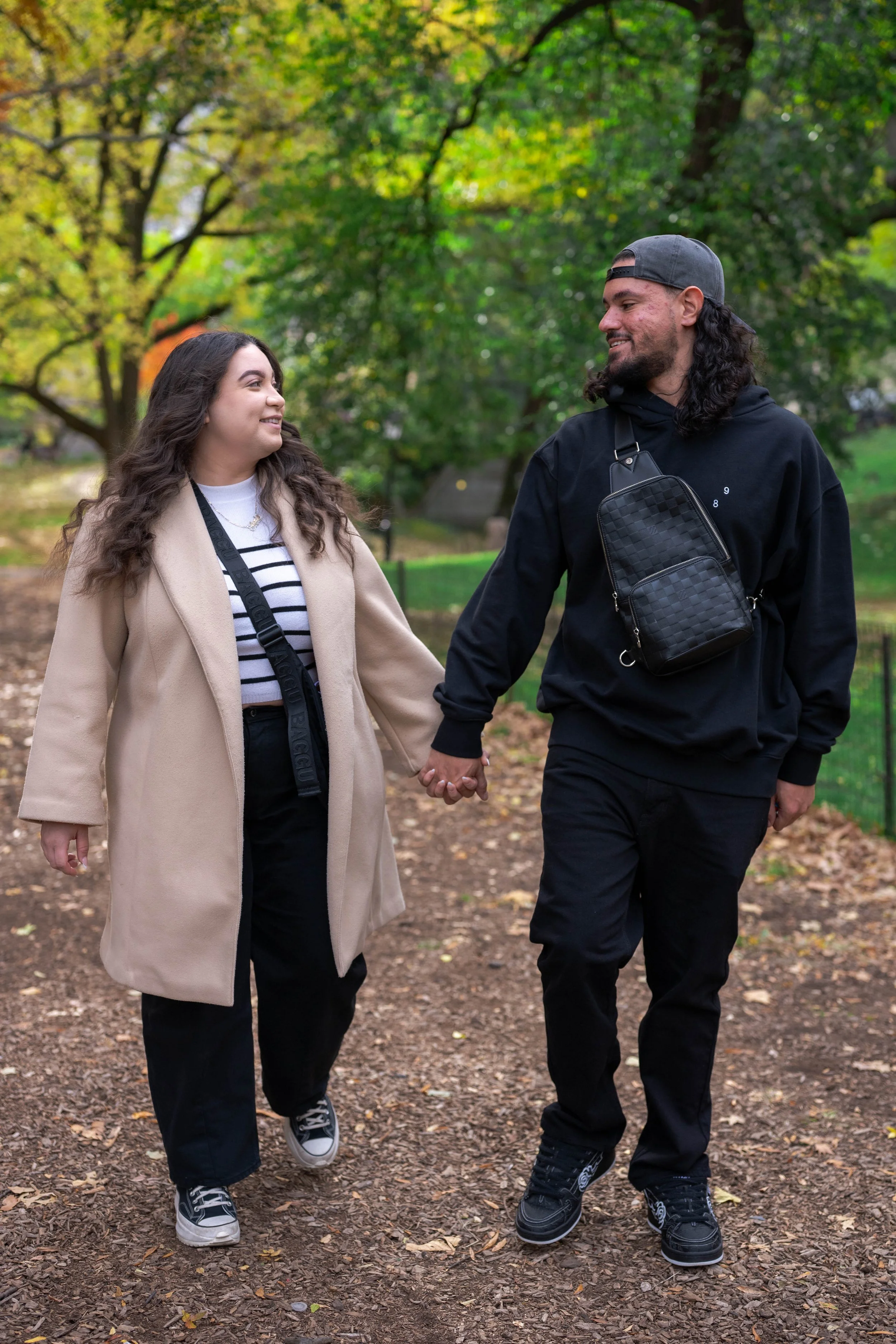 A young couple holding hands and walking in a park with green trees, smiling at each other.