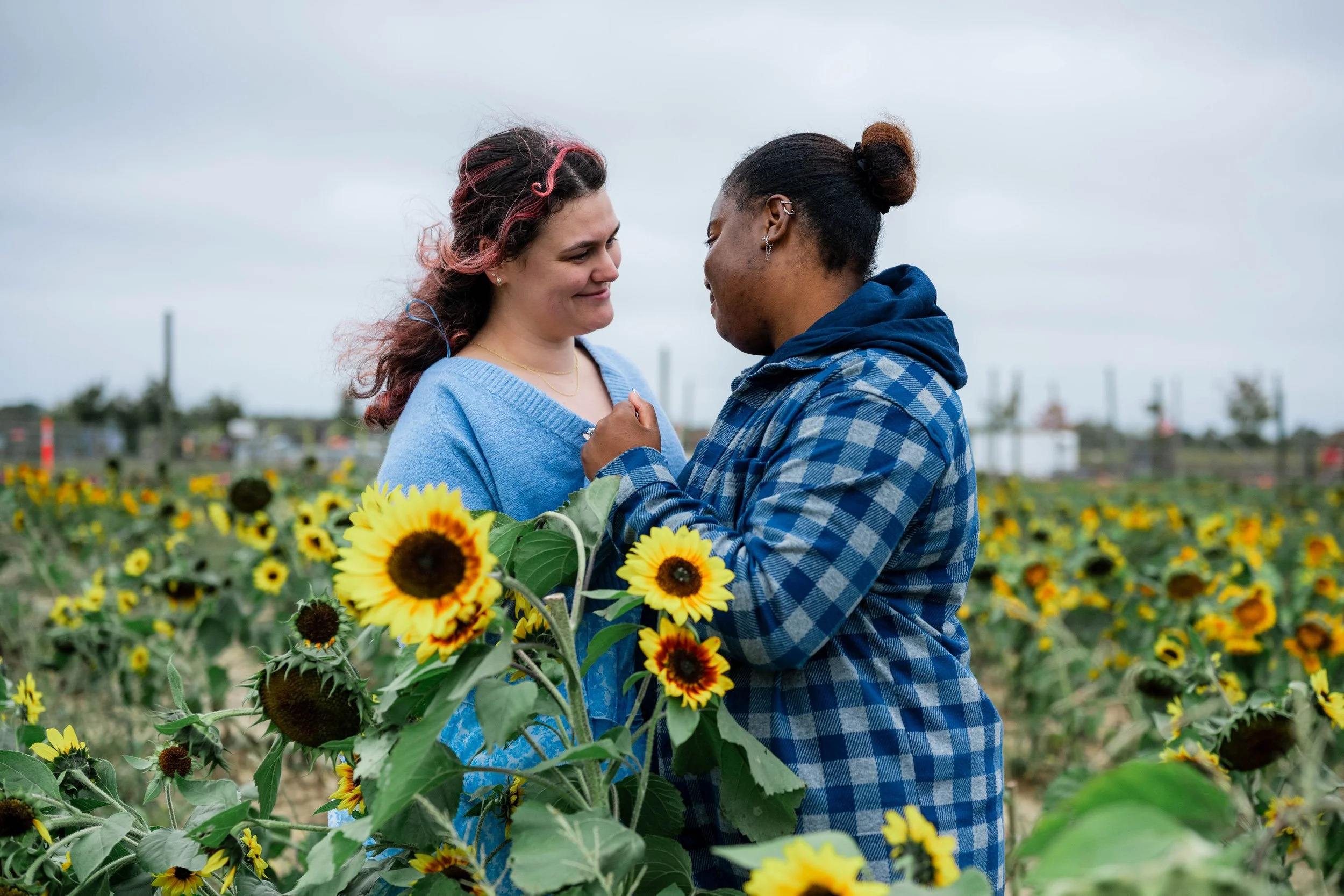 Two women standing close together in a sunflower field, smiling at each other, with overcast sky and industrial structures in the background.