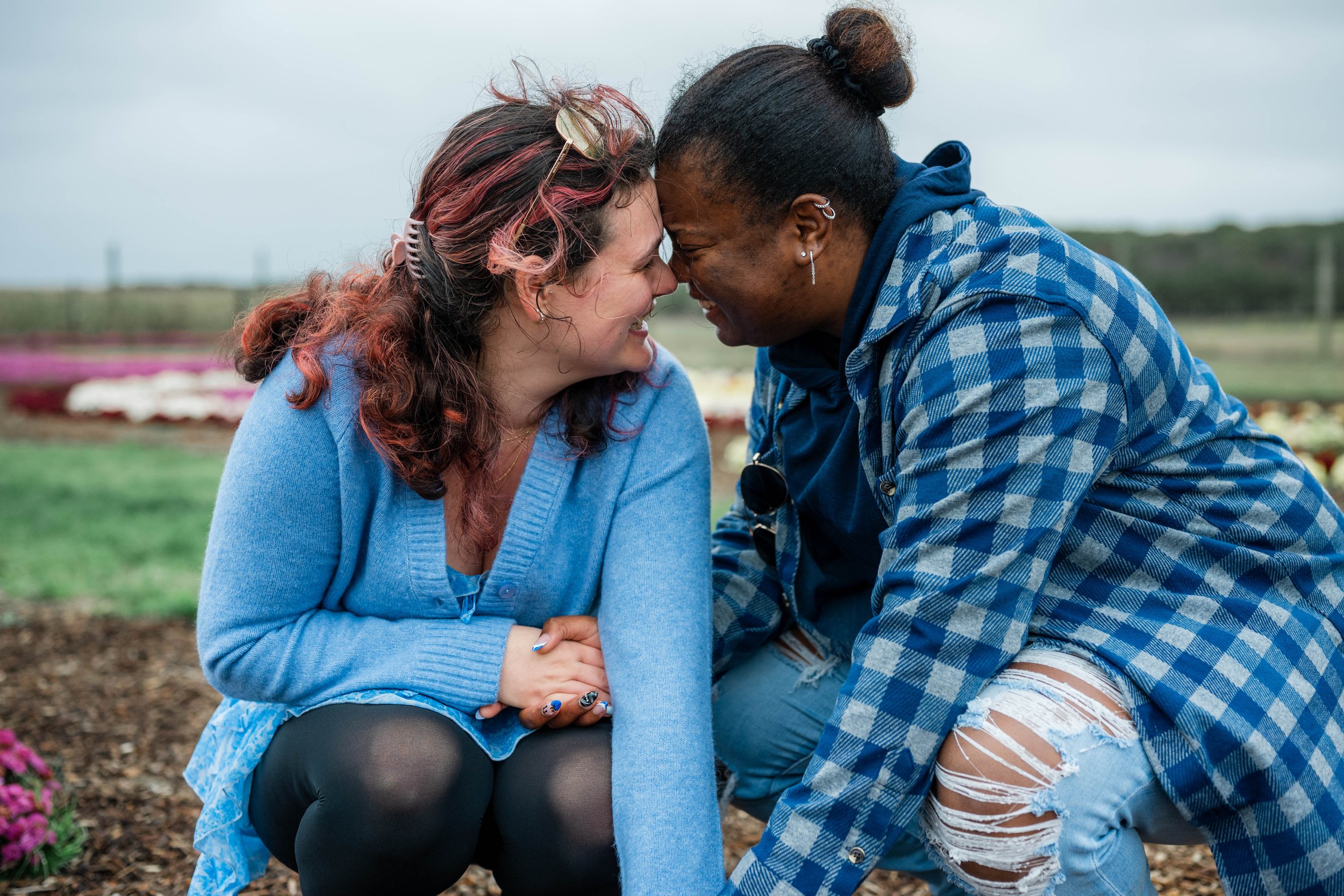 Two women in a garden, touching foreheads and smiling at each other, holding hands.