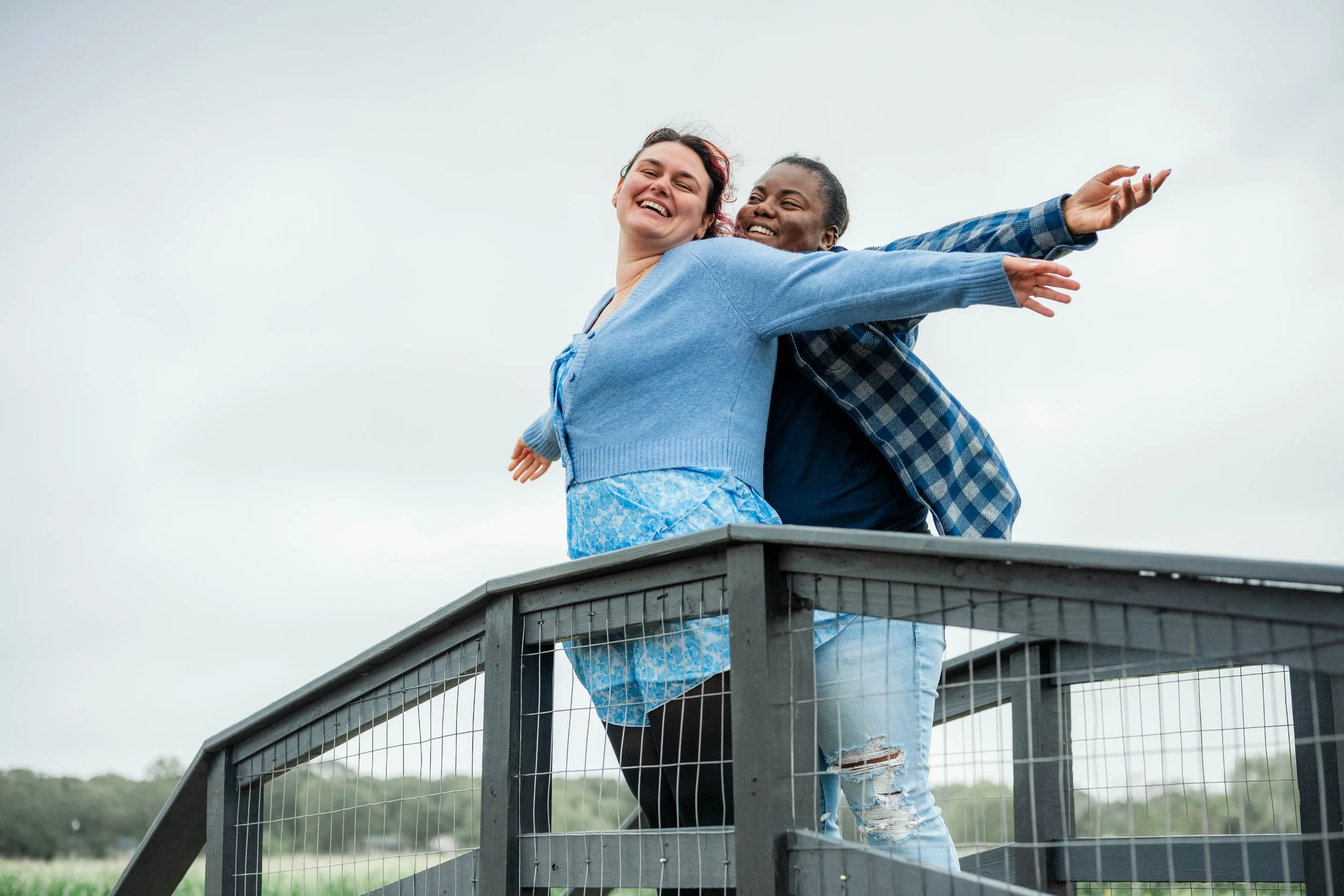 Two women stand on a small outdoor platform with arms outstretched, smiling and enjoying the moment against a cloudy sky background.