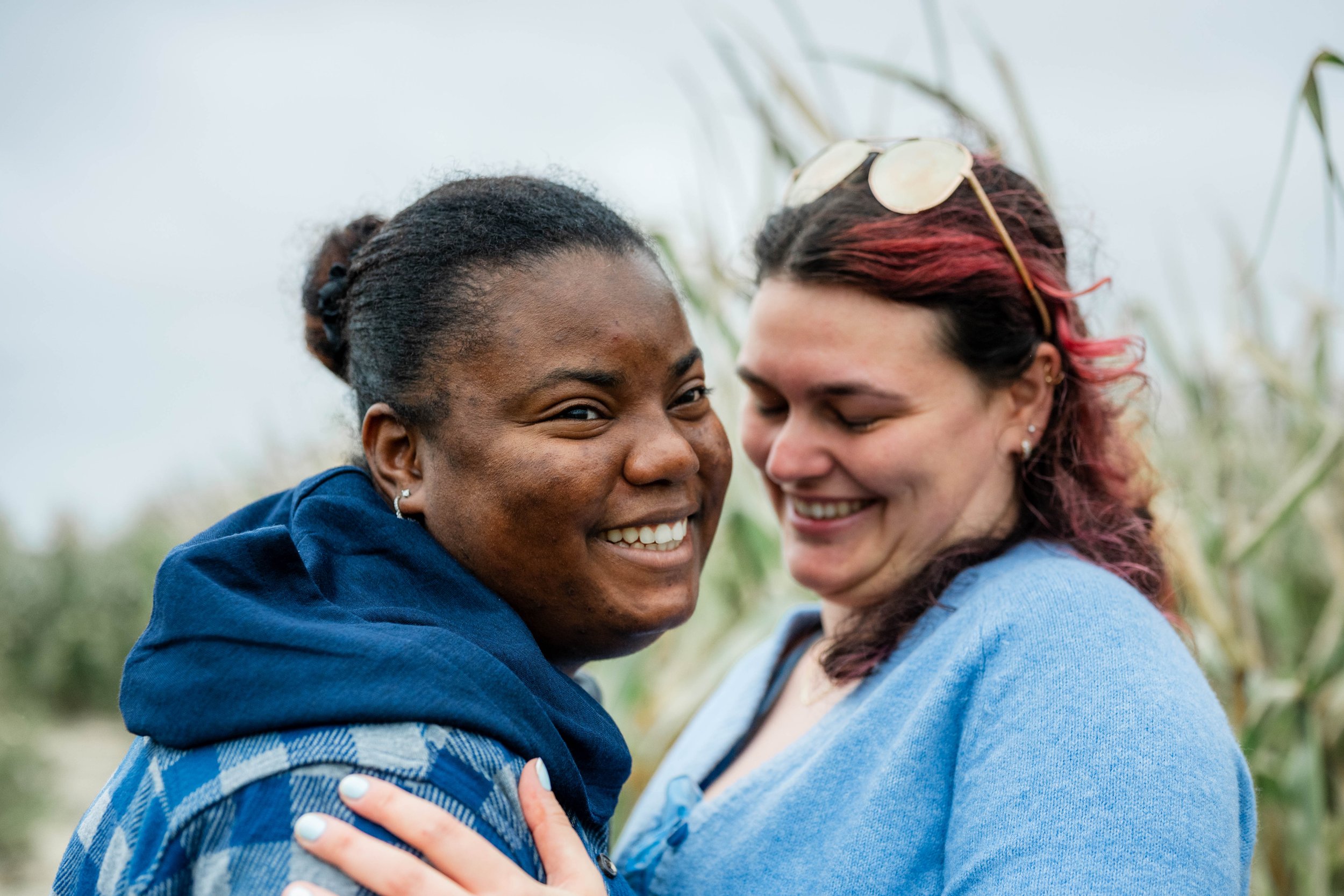 Two women smiling and hugging outdoors, with tall grass or crops in the background.