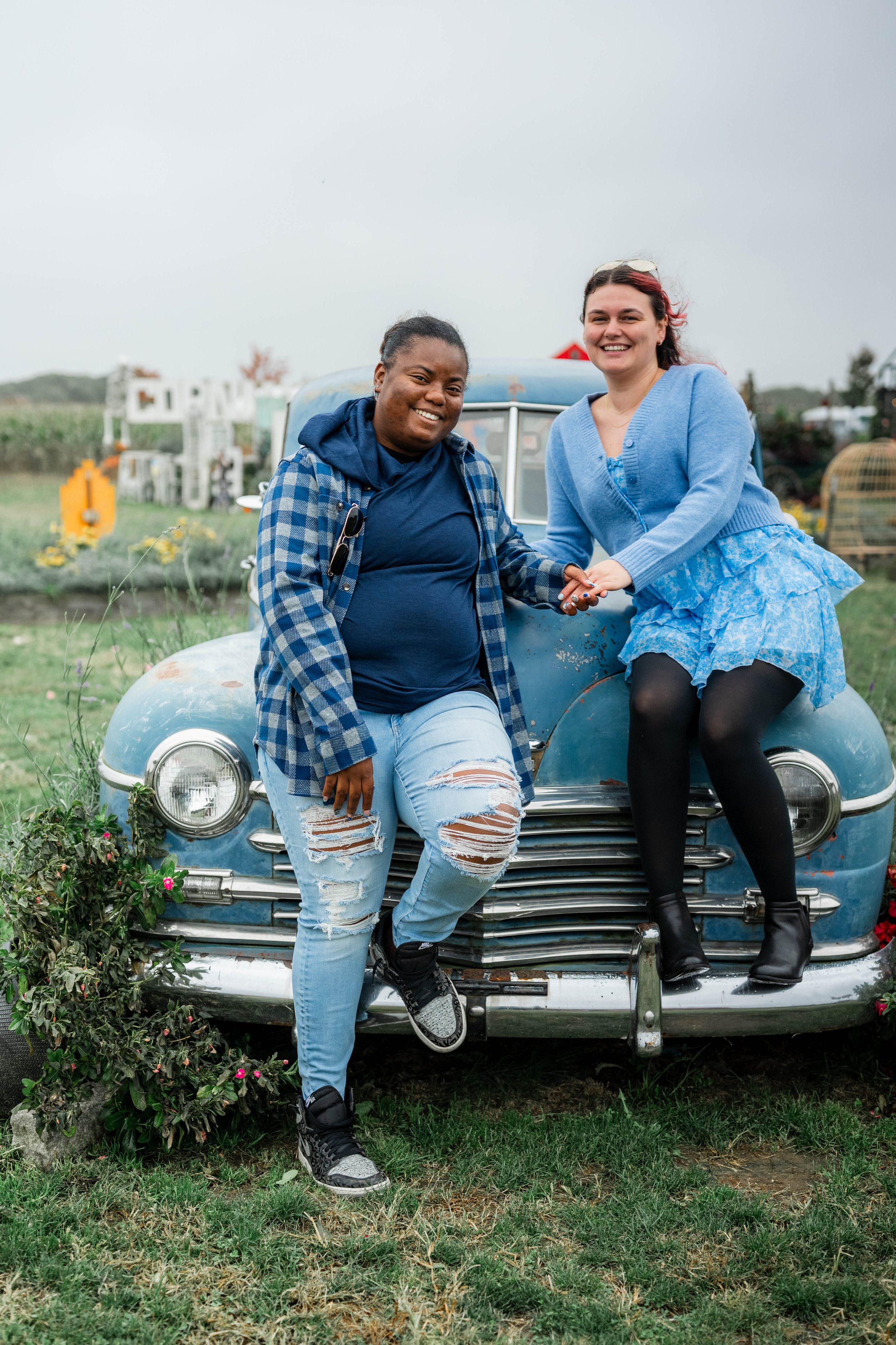 Two women smiling and holding hands, one sitting on the hood of a vintage blue car and the other standing with one foot on the car, outdoors in a garden or field.