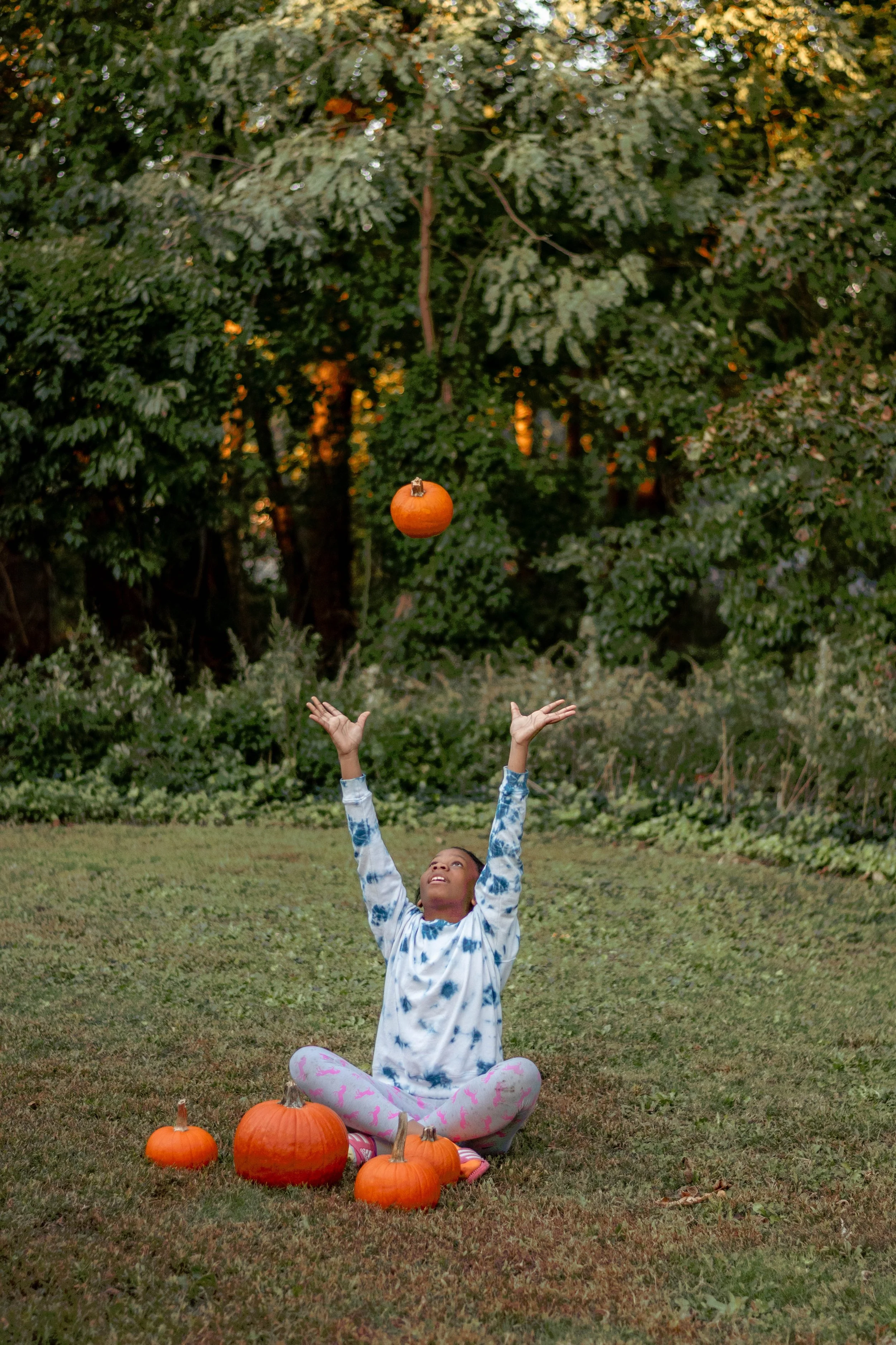 A young girl sitting on the grass in a park surrounded by pumpkins, throwing a pumpkin into the air, with trees in the background.