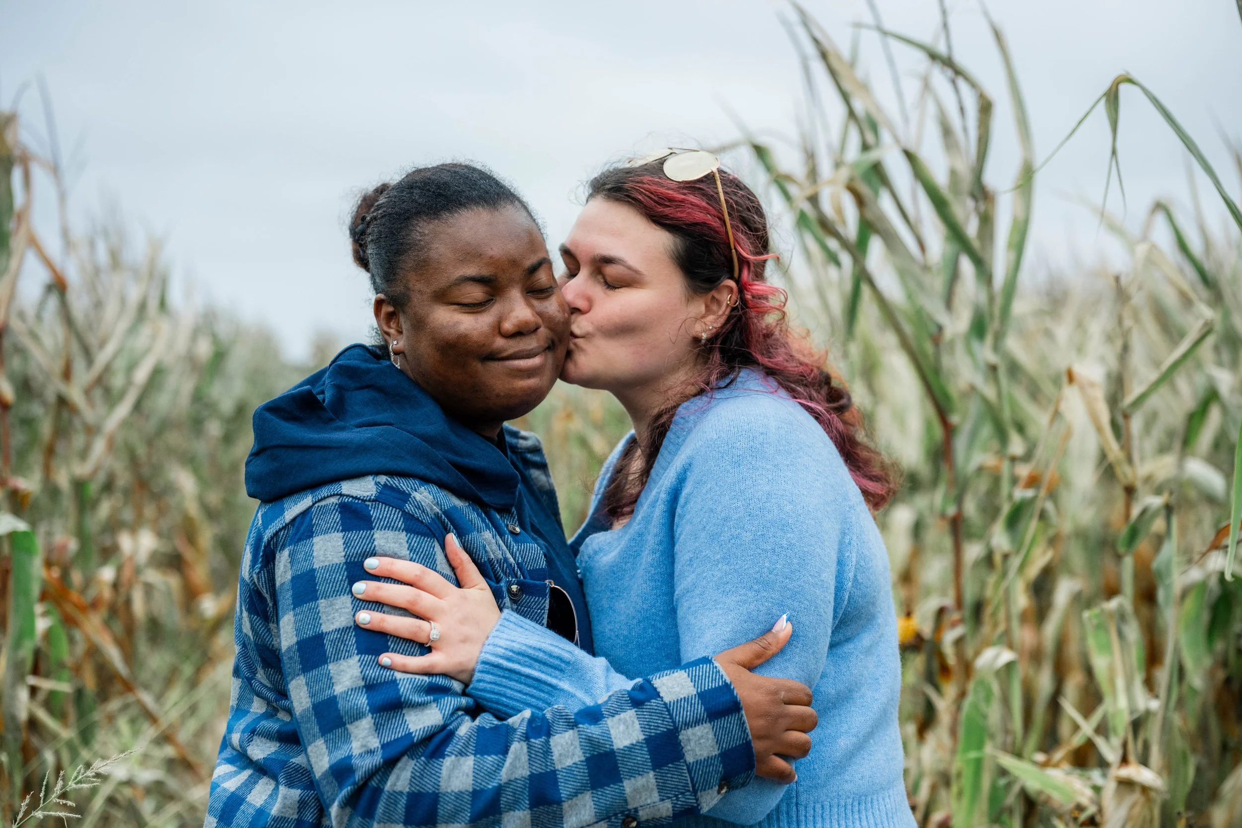 Two women embracing and sharing a kiss in a cornfield, with cloudy sky in the background.