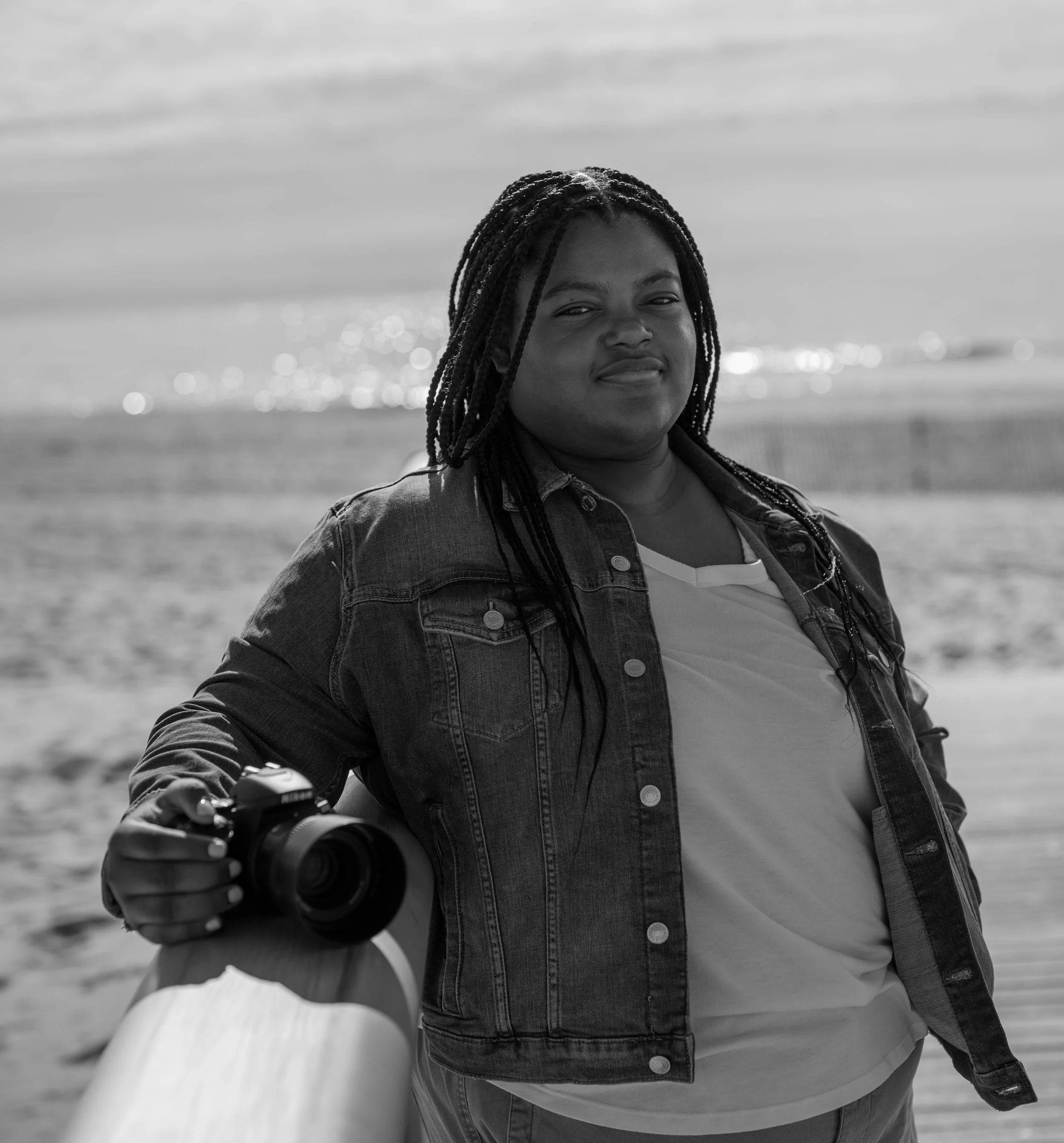 A woman standing on a beach holding a camera, wearing a denim jacket and looking at the camera.