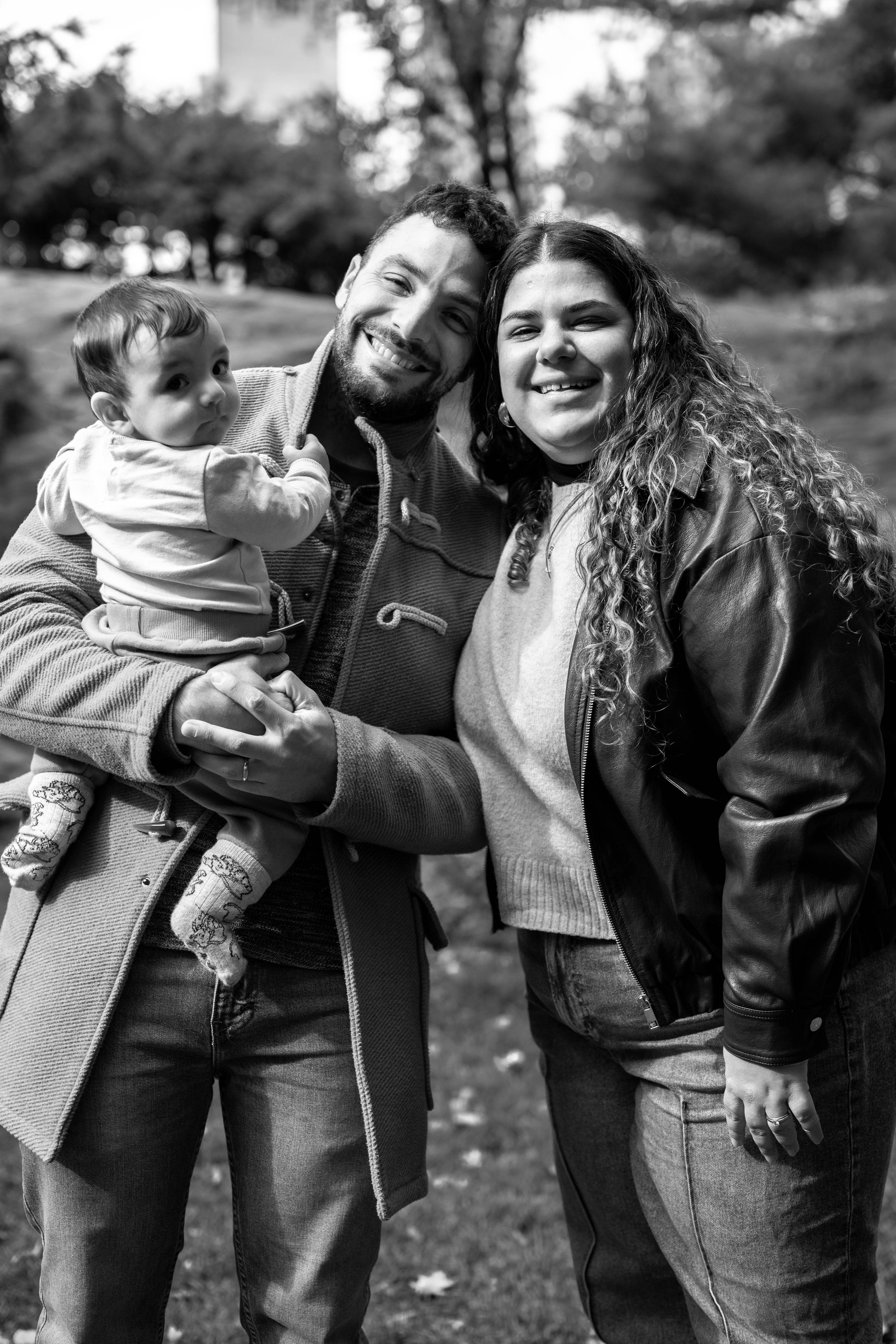 A family of three outdoors, smiling for a photo. The father holds a baby girl, and a woman stands beside them. Trees and blurred landscape in the background.