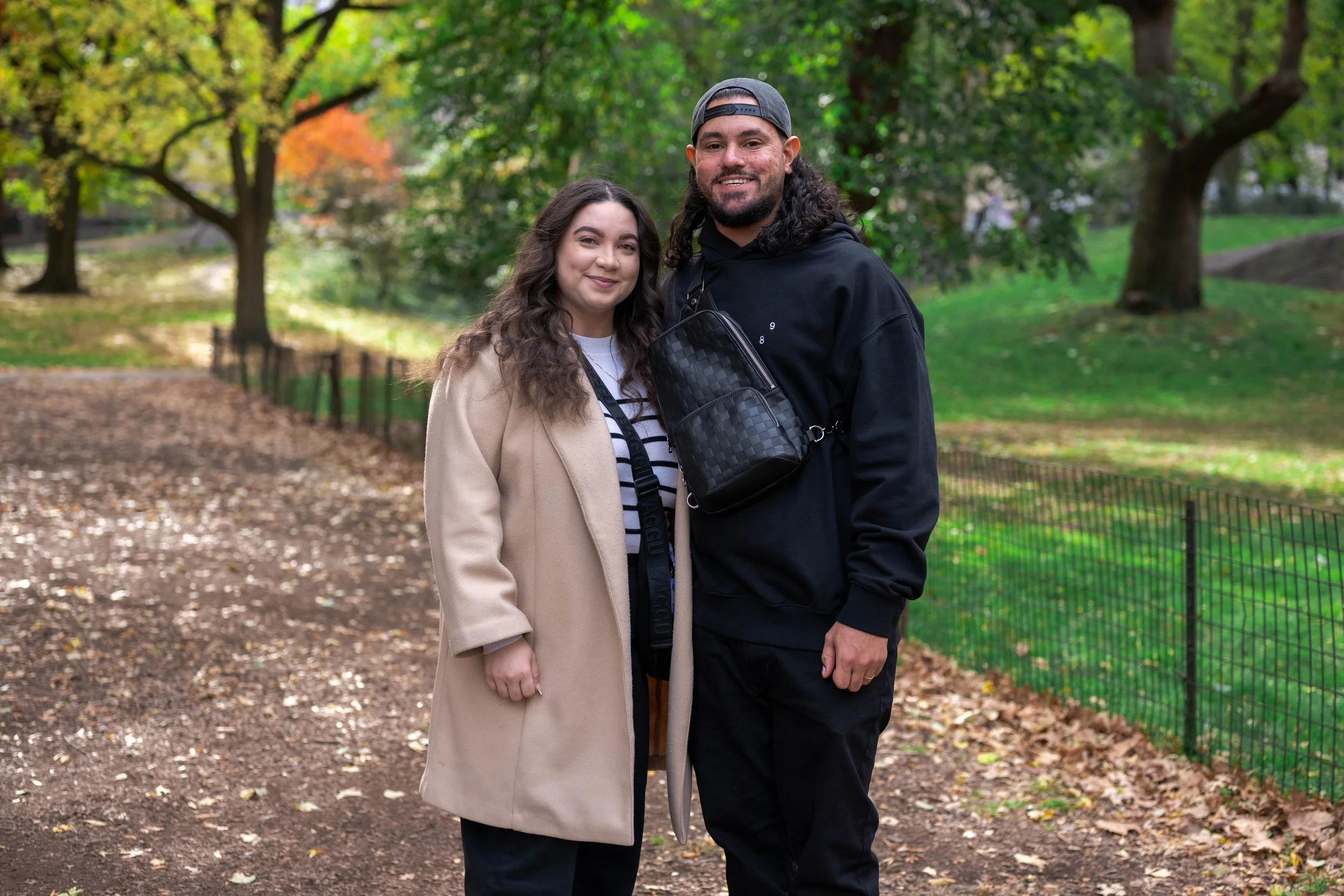 A woman and a man standing together outdoors in a park with trees and fallen leaves, smiling at the camera.