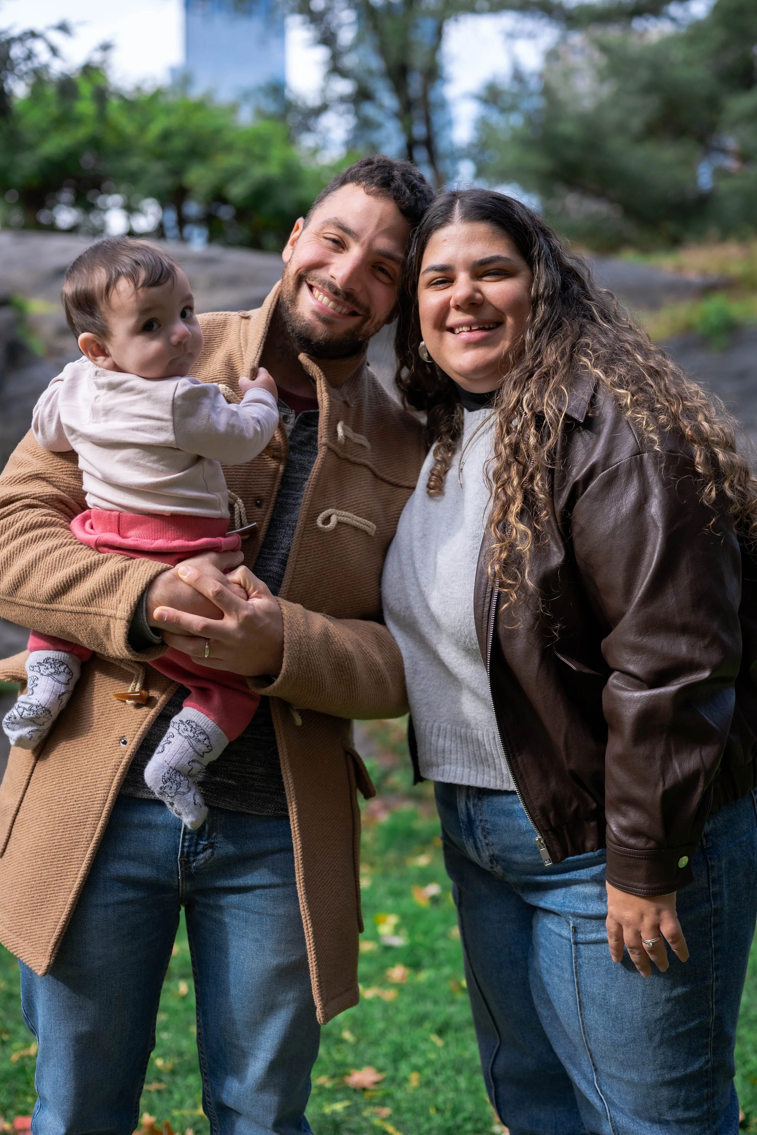 A smiling family of three outdoors, with a man holding a baby girl and a woman standing beside them, in a park with trees and grass.