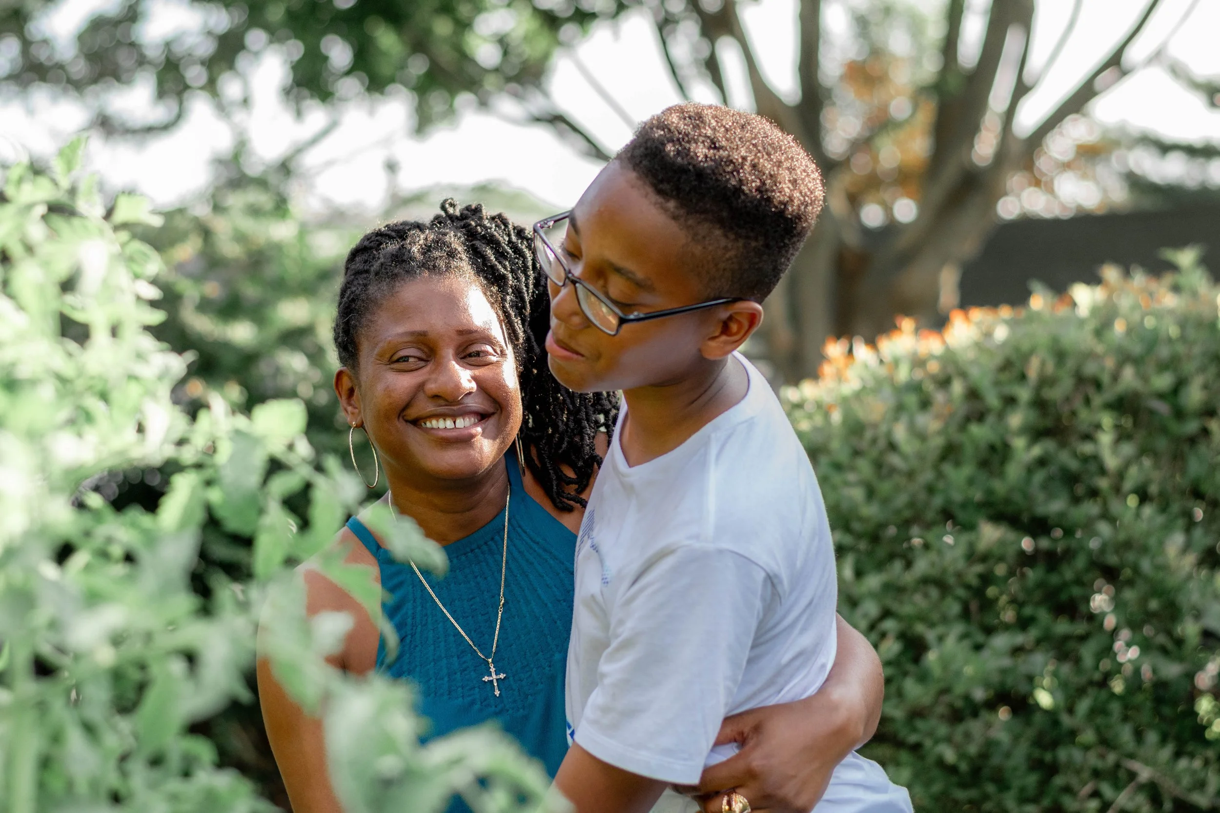 A woman with dark skin and dreadlocks smiling and hugging a young man with glasses and dark skin in a white shirt outdoors surrounded by green foliage.