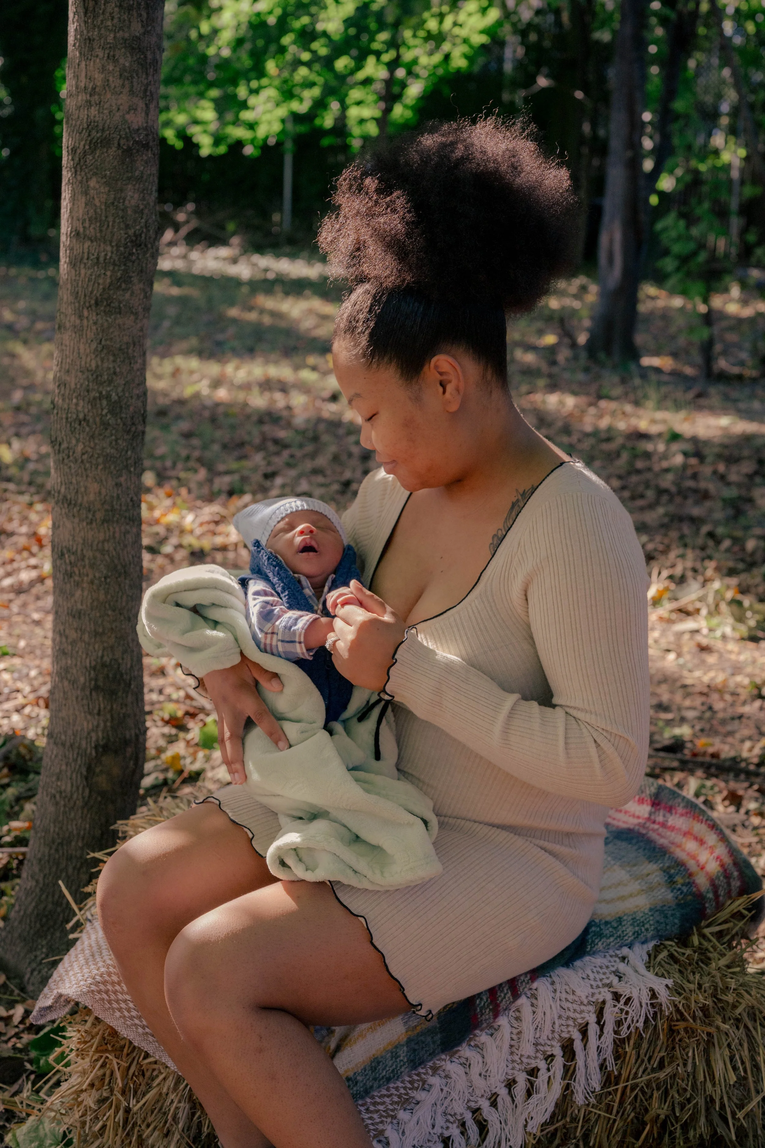 A woman with natural hair holding a newborn baby in a forest setting, sitting on a hay bale with a blanket, both looking at each other.