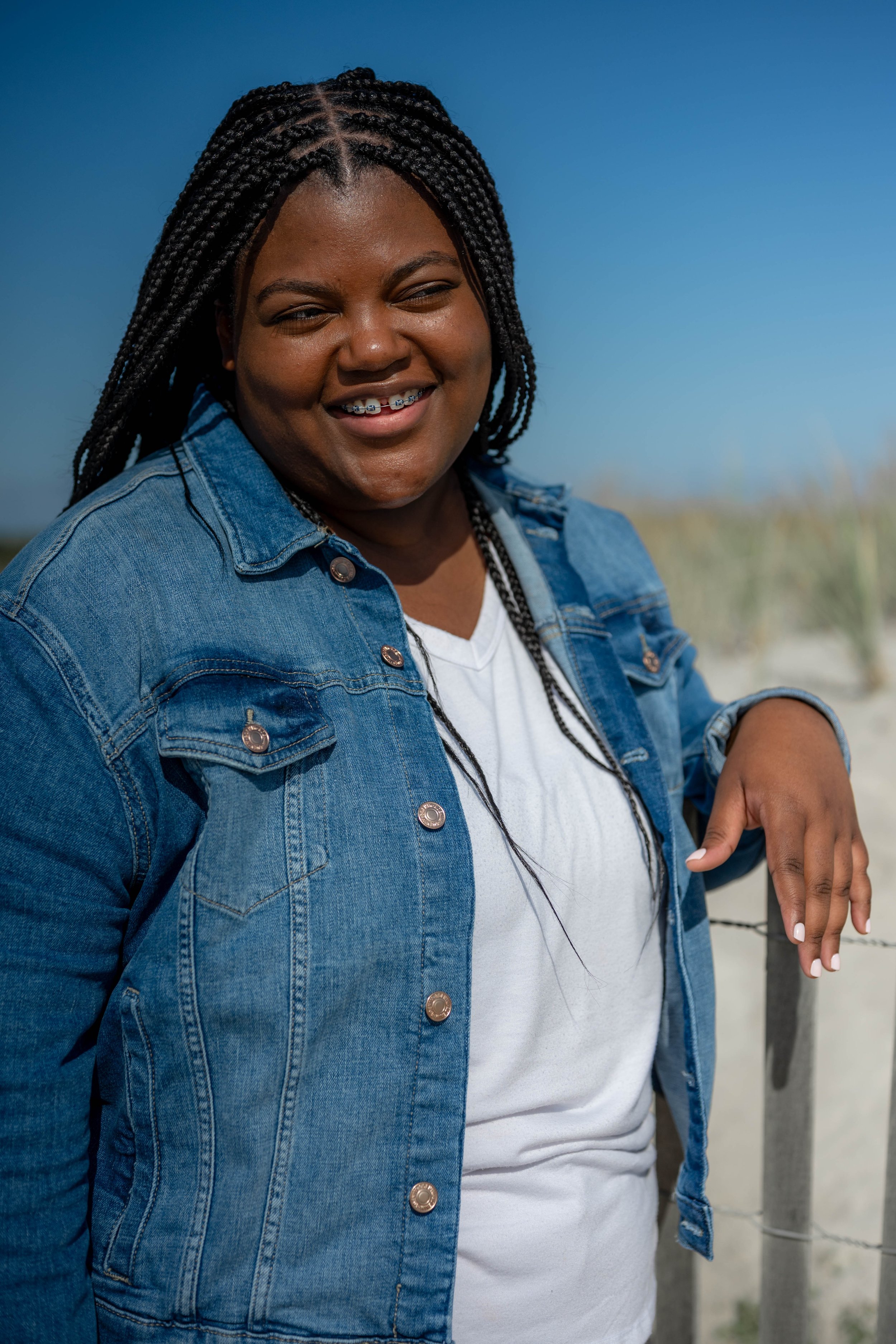 A smiling woman with braids wearing a denim jacket and white T-shirt standing outdoors on a sunny day, with a blue sky and beach in the background.