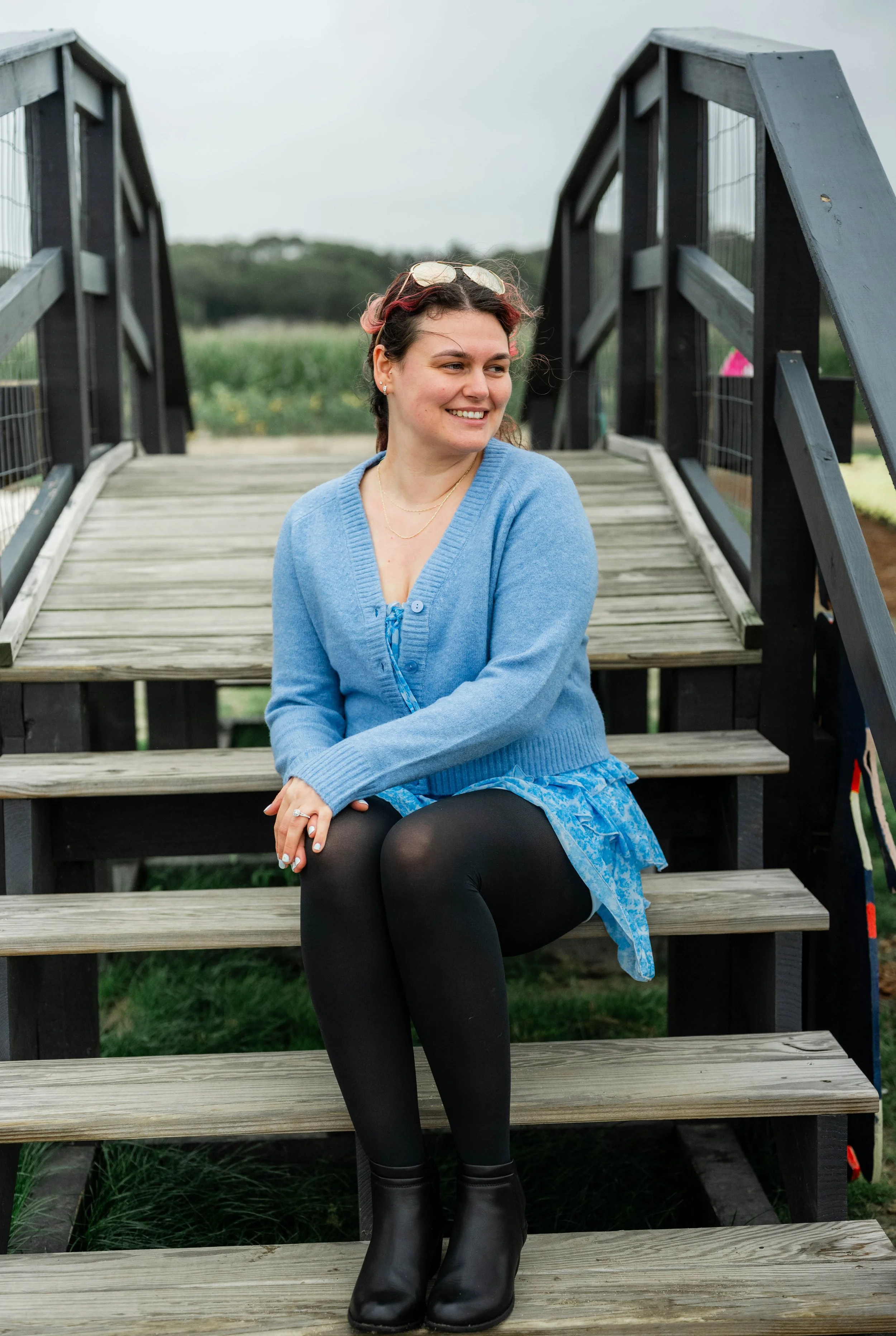 A woman with curly hair sitting on wooden stairs outdoors, smiling and looking to the side, wearing a blue cardigan, black tights, and black ankle boots, with sunglasses on her head.