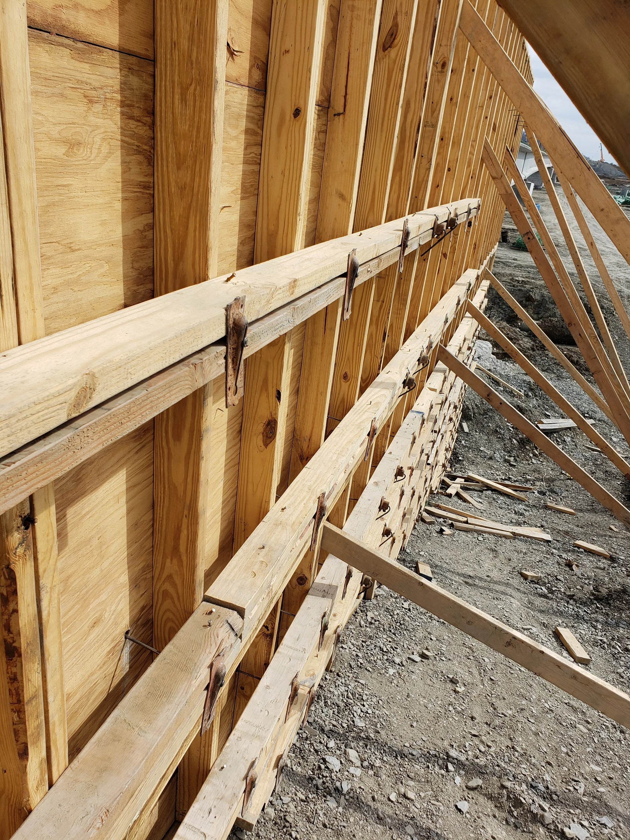 Close-up view of wooden framing work on a construction site Tulsa, showing vertical and horizontal wooden beams and supports, with some metal fasteners and brackets, and dirt ground below.