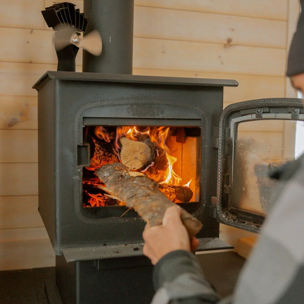 Personne alimentant le poêle à bois de l’éco-chalet Ontara, un bâtiment autonome en énergie favorisant chaleur et convivialité.