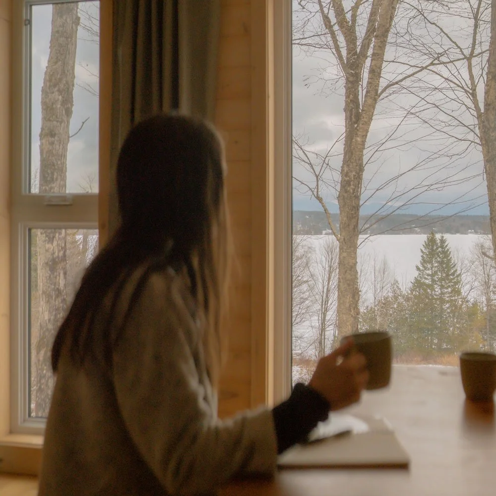 Femme regardant par une grande fenêtre, tasse à la main, face à un paysage enneigé du Québec