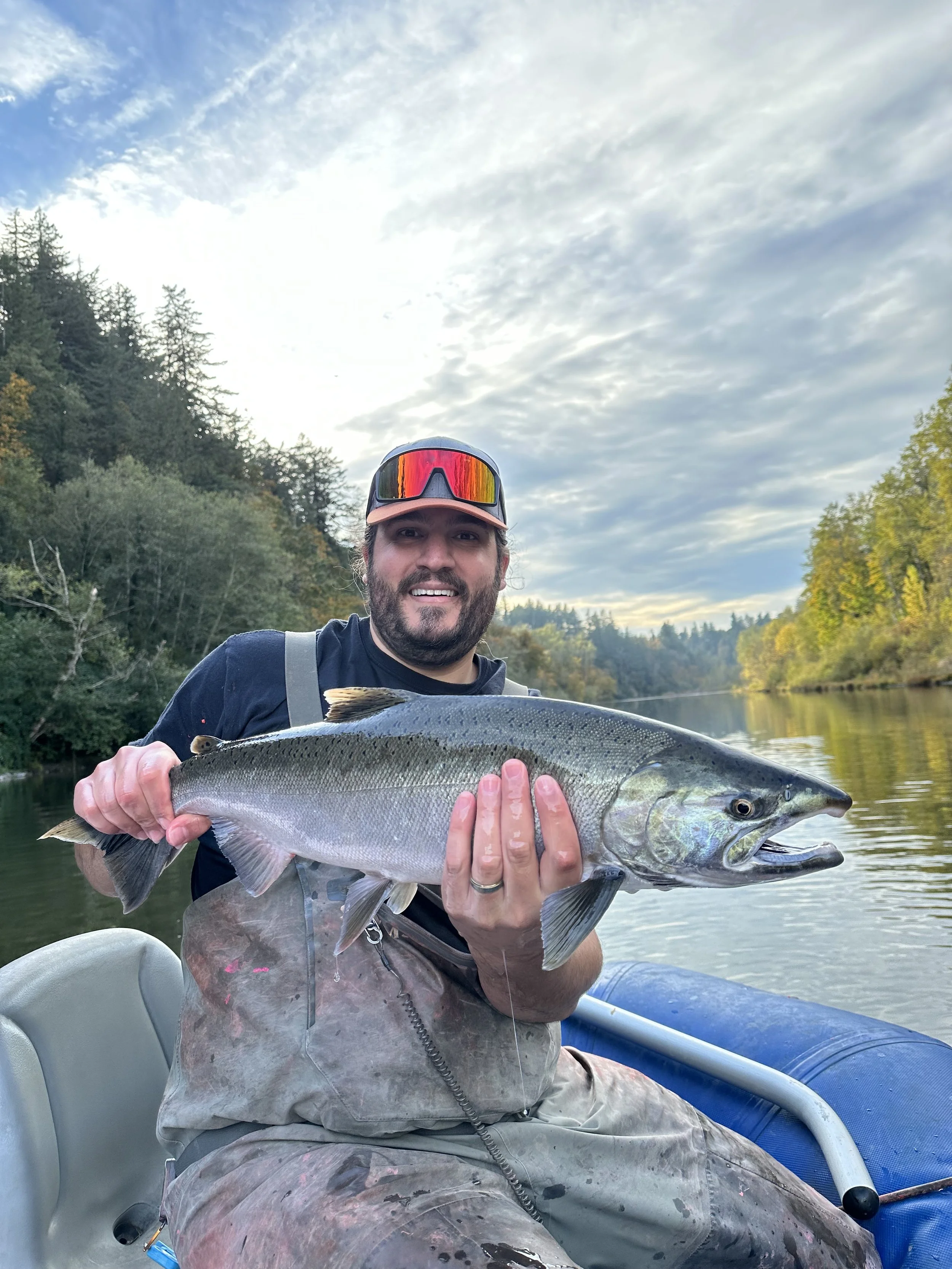 A man in sunglasses and fishing gear holding a large fish on a boat with a river and forested landscape in the background.