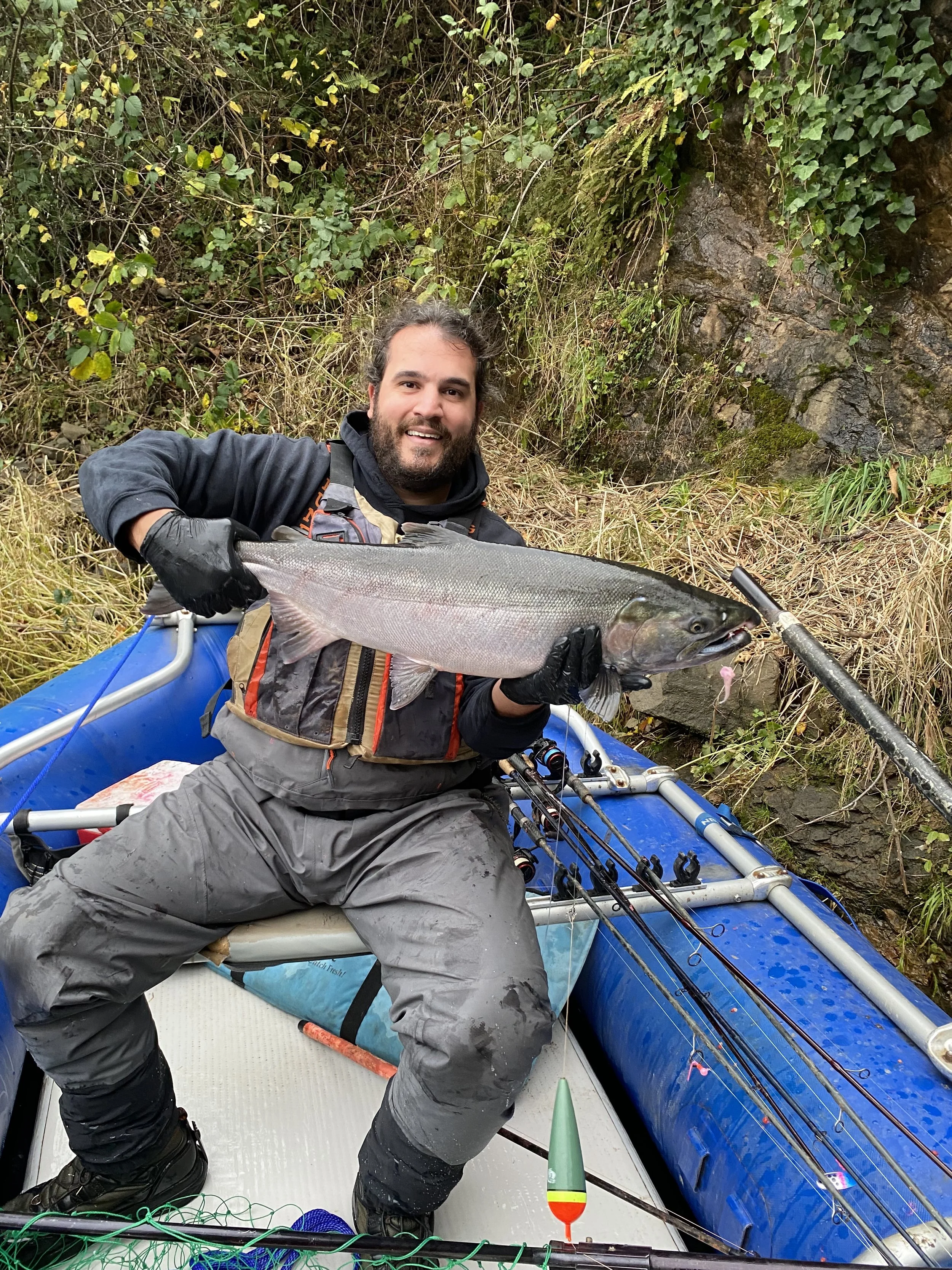 A man with a beard, smiling, sitting in a small blue inflatable boat, holding a large fish, possibly a salmon, with a pink fishing lure in its mouth, near a rocky and leafy riverside.