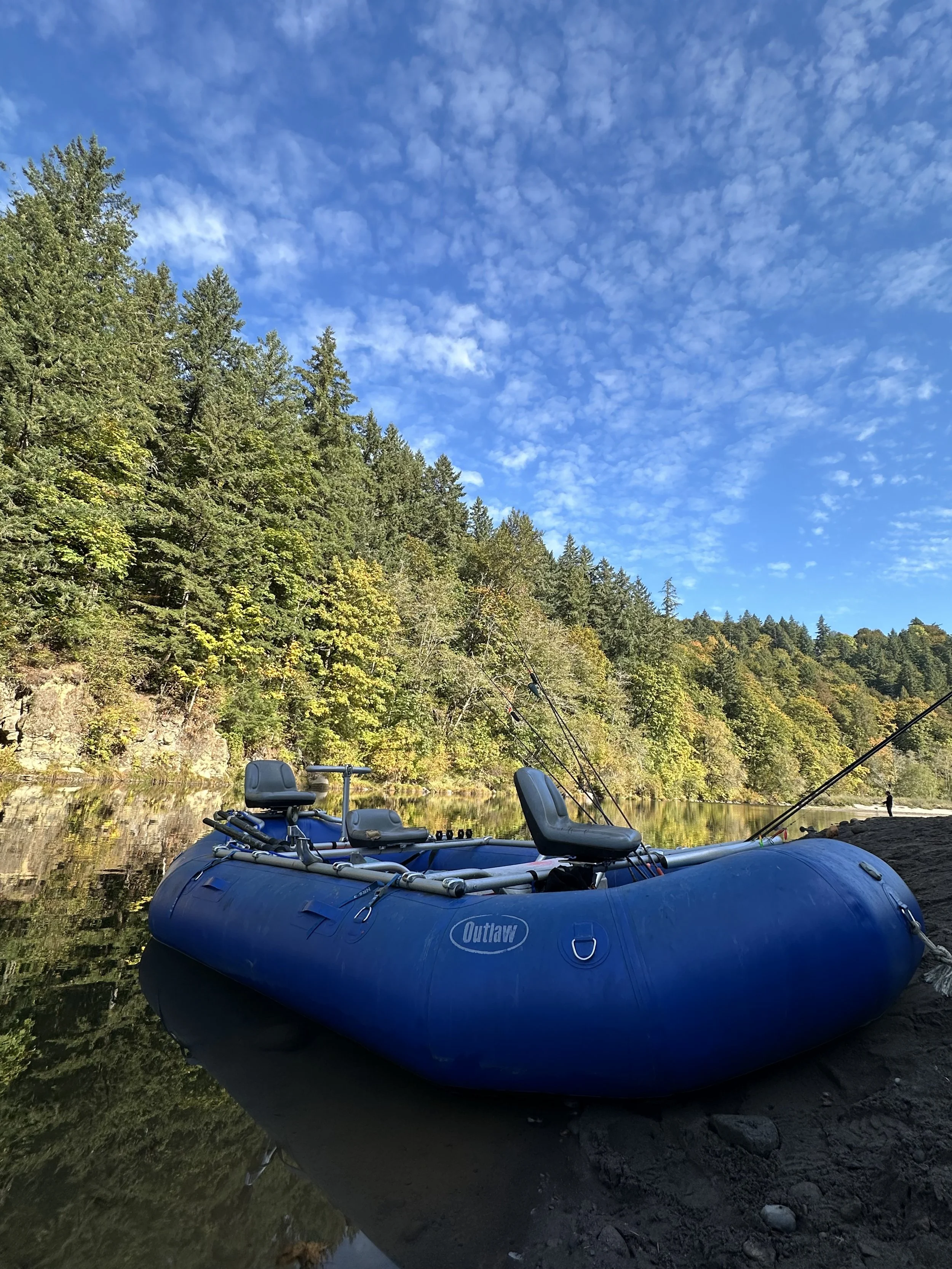 A blue inflatable boat with fishing rods is docked on a rocky shore beside a calm river, with a forested hillside and a partly cloudy blue sky in the background.