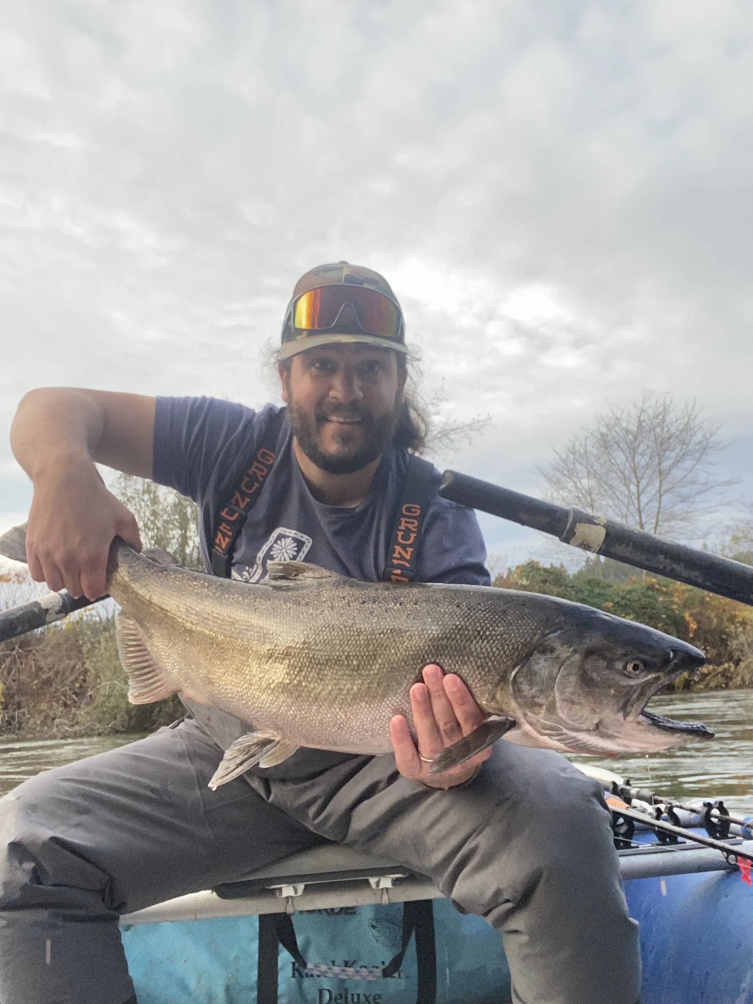 Man in fishing gear holding large fish on a boat with fishing rods, trees and cloudy sky in the background.
