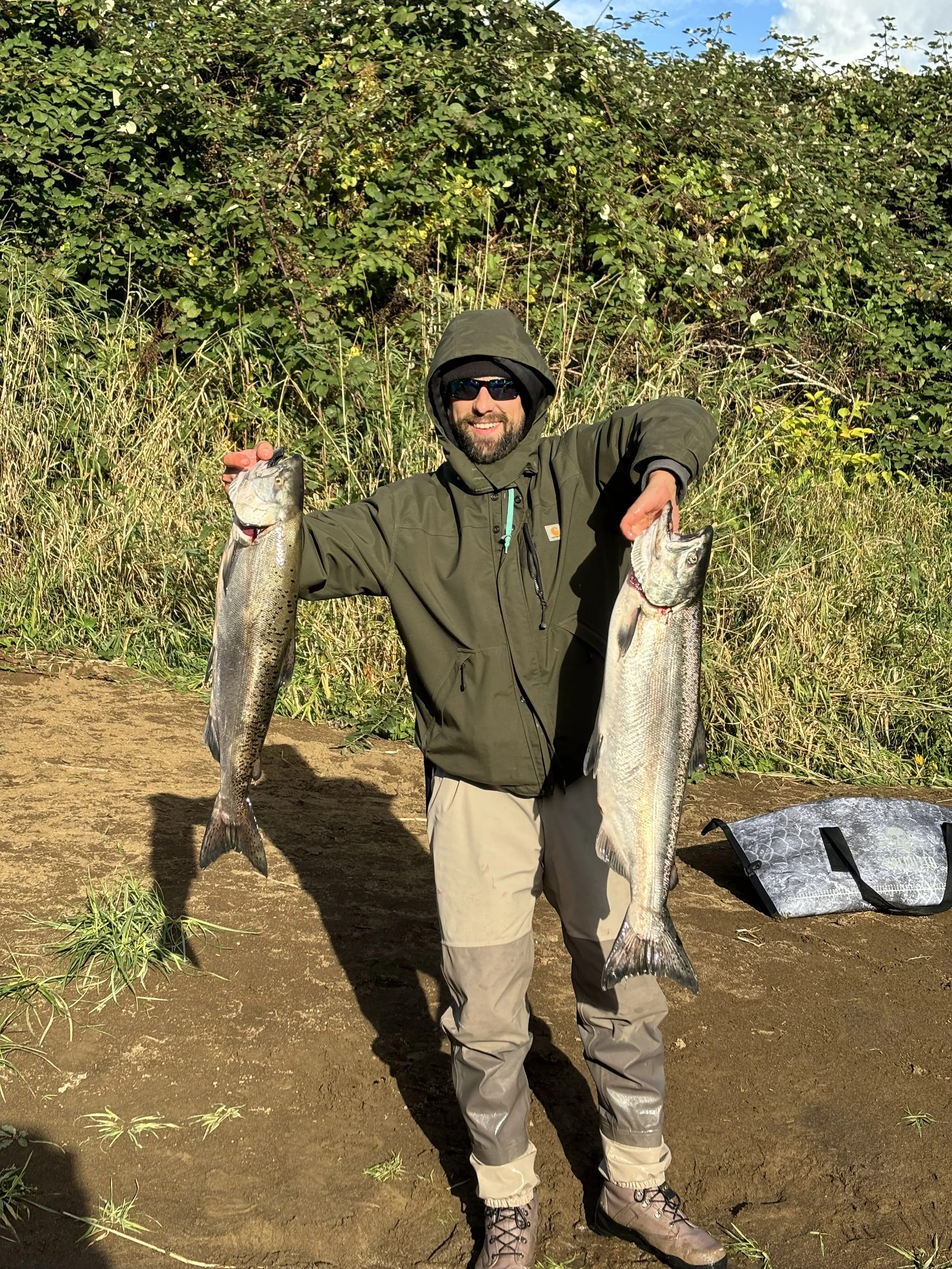 A man wearing sunglasses and a hooded jacket holding two large fish, one in each hand, on a dirt path near tall grass and greenery.