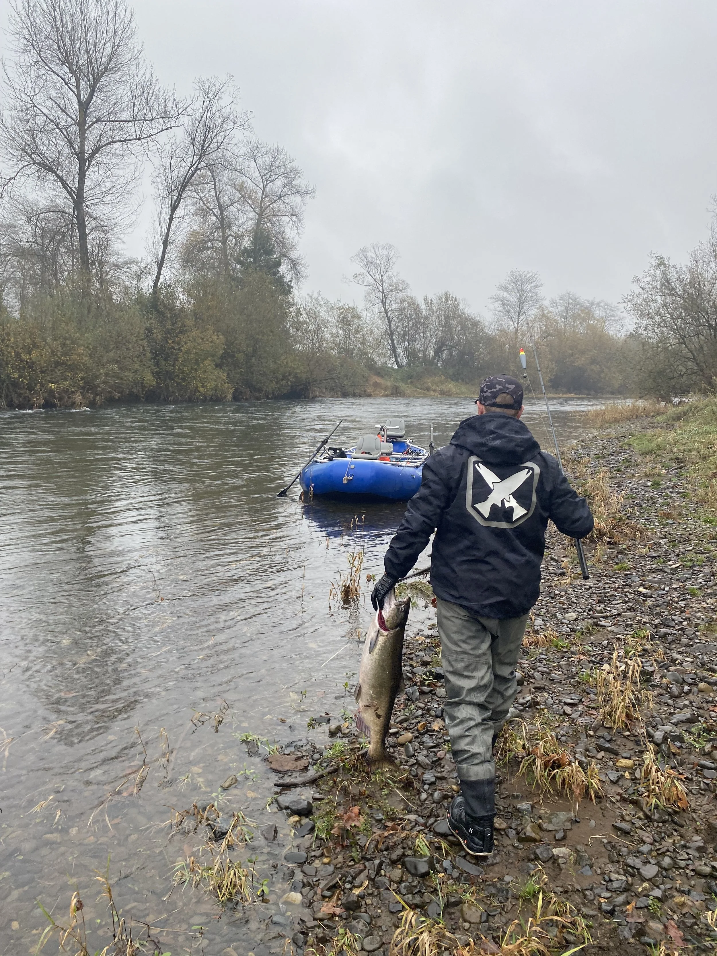 Man walking on rocky riverbank holding a large fish, with a fishing boat anchored nearby on a cloudy day.