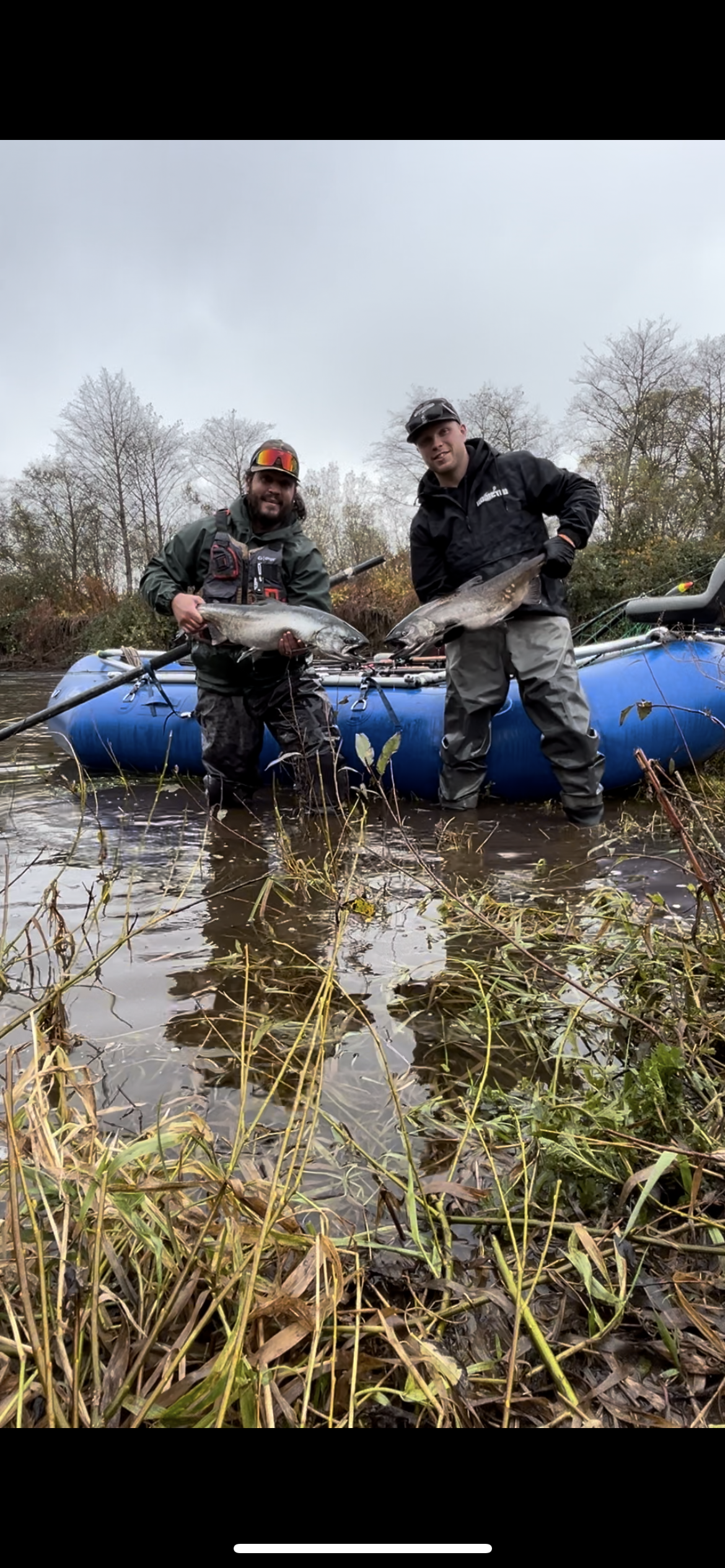 Two men standing in a shallow river holding large fish, with a blue inflatable boat and trees in the background.
