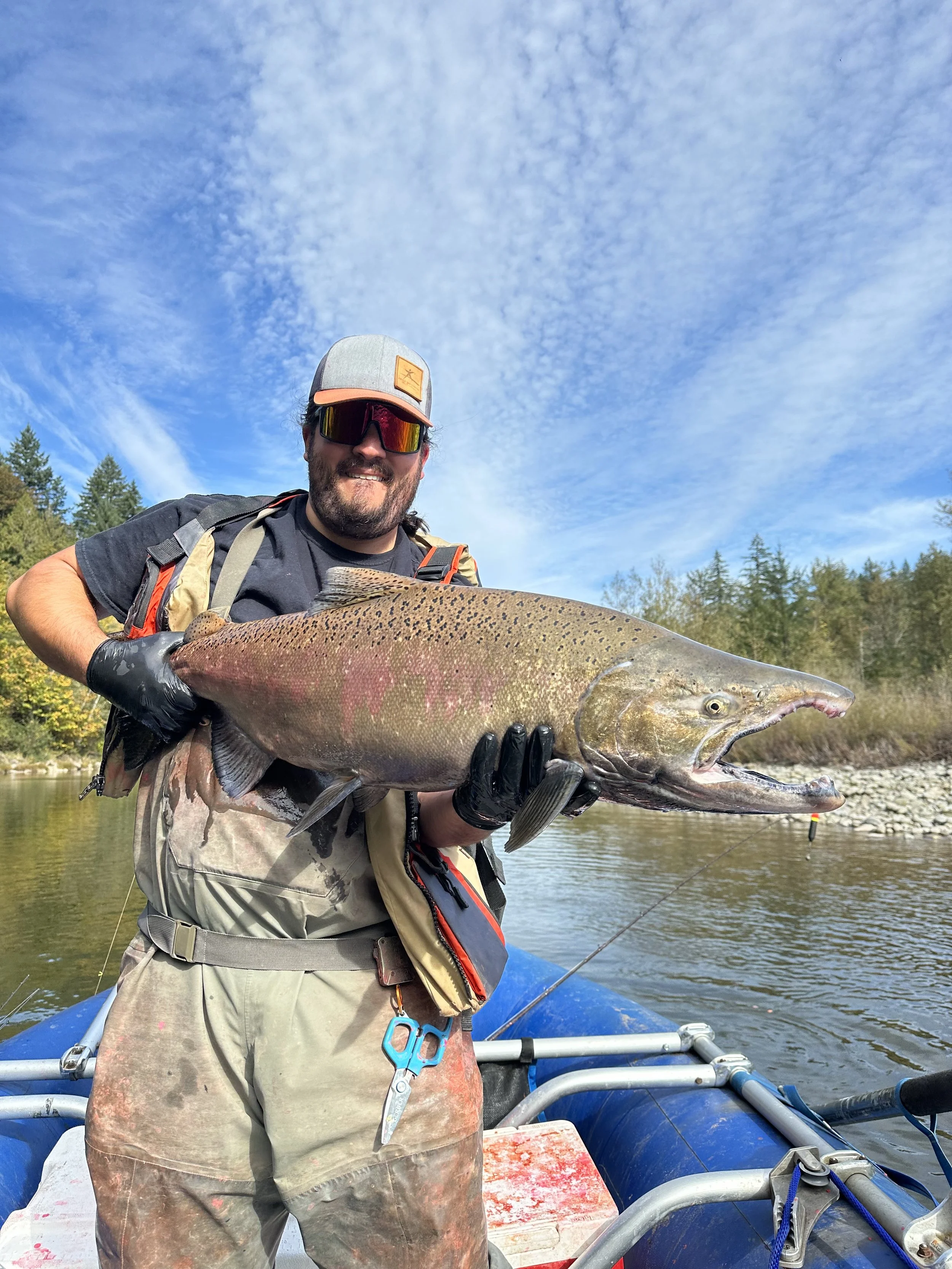 A man wearing sunglasses and a cap holding a large fish on a boat in a river, with trees and blue sky in the background.