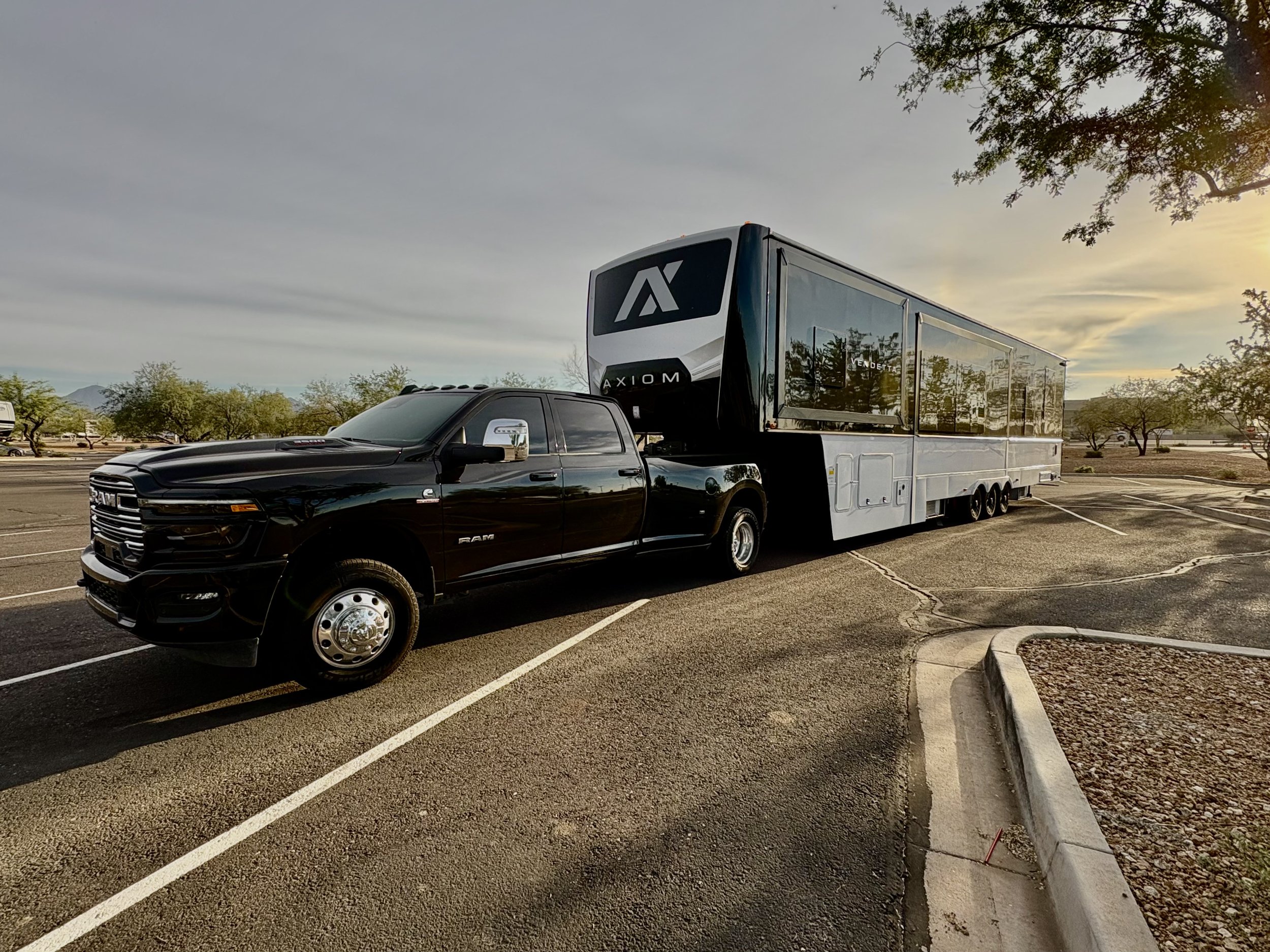 Black pickup truck towing a large modern trailer in a parking lot during sunset, with trees and a cloudy sky in the background.
