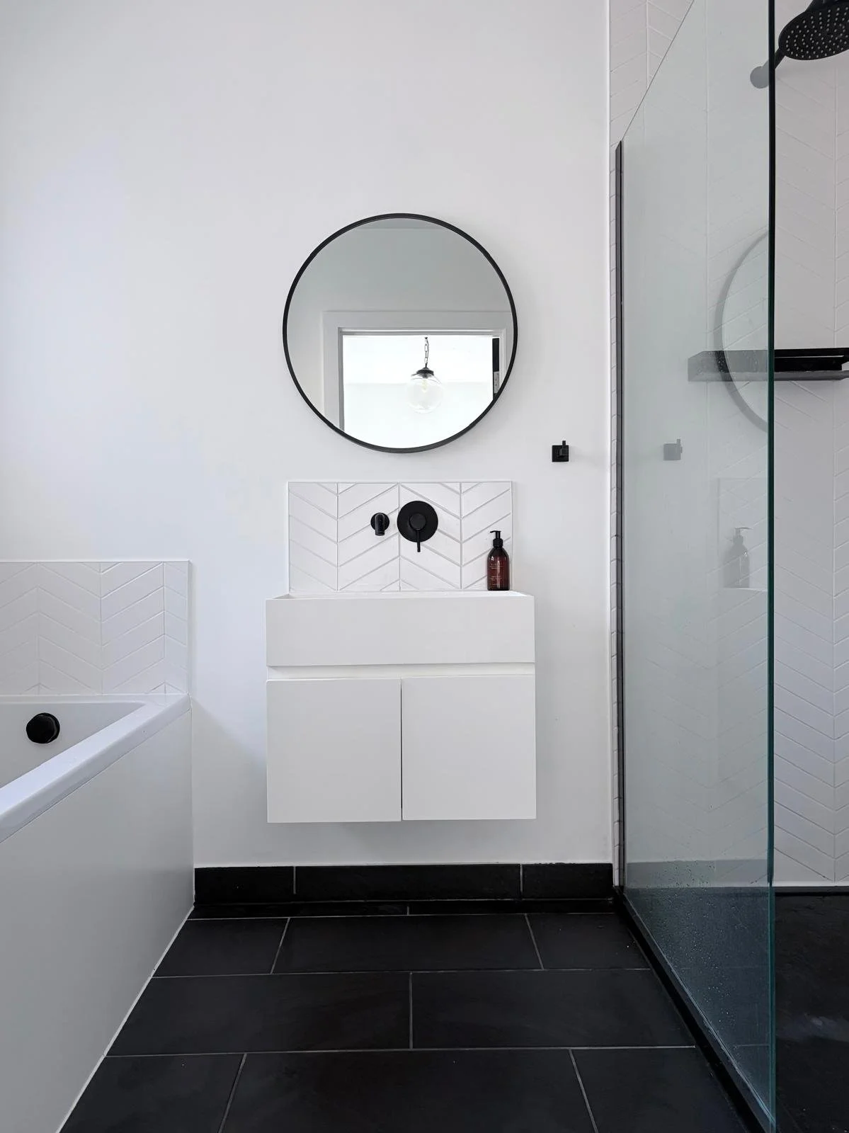 Modern bathroom with a freestanding bathtub, green tiled accent wall, terrazzo lower wall and floor, and a glass-enclosed shower area with grey vertical tiles. There is a frosted window with two bottles on the window sill.