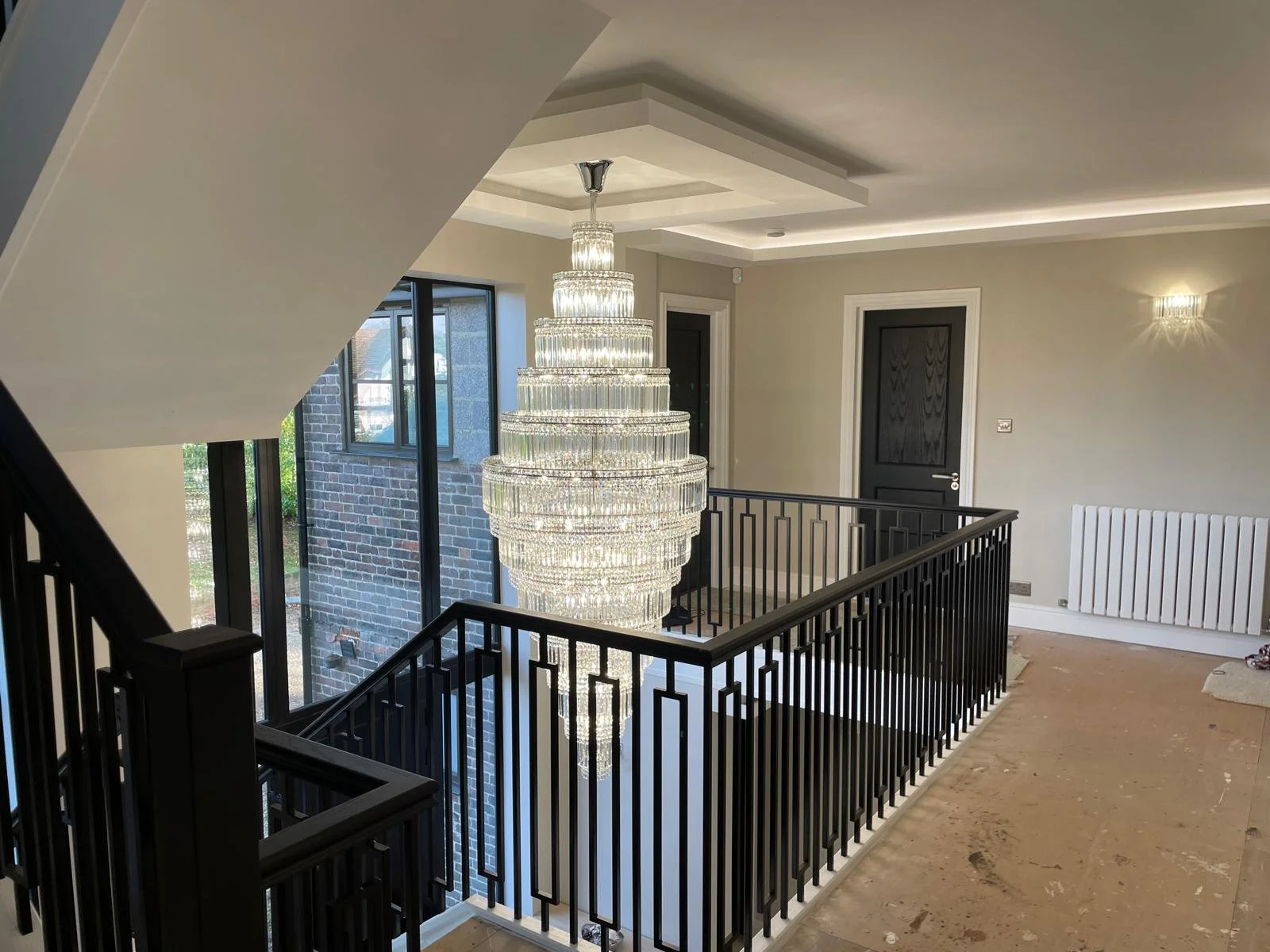 Modern interior space featuring a grand crystal chandelier, black staircase railing, large window with brick exterior view, light-colored walls, and a black door.