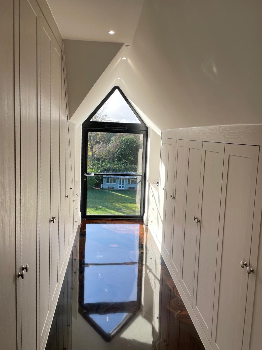 A narrow hallway with cabinets on both sides, leading to a glass door with a triangular window at the top, showing a view of a grassy yard with trees and a small building outside.