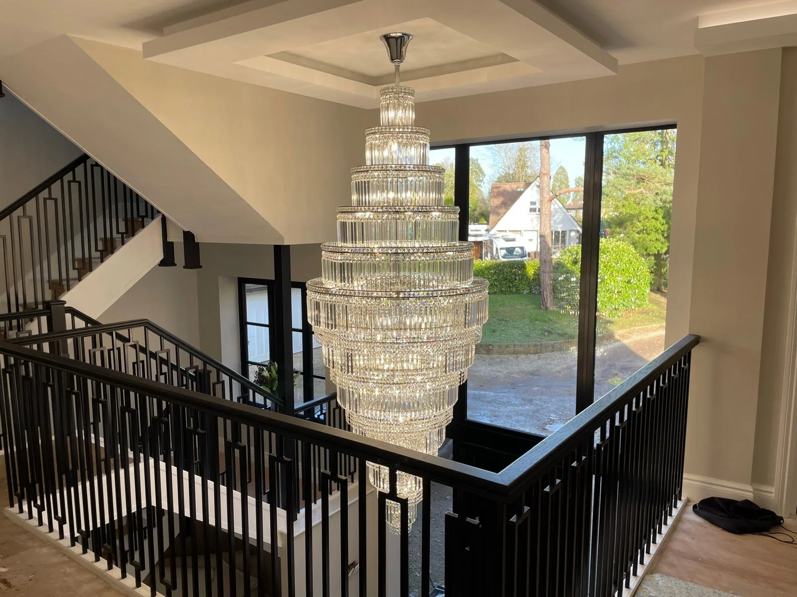 A large, multi-tiered crystal chandelier hanging above a staircase with black railing inside a modern home, with a large window showing an outdoor view of green trees and a neighboring house.
