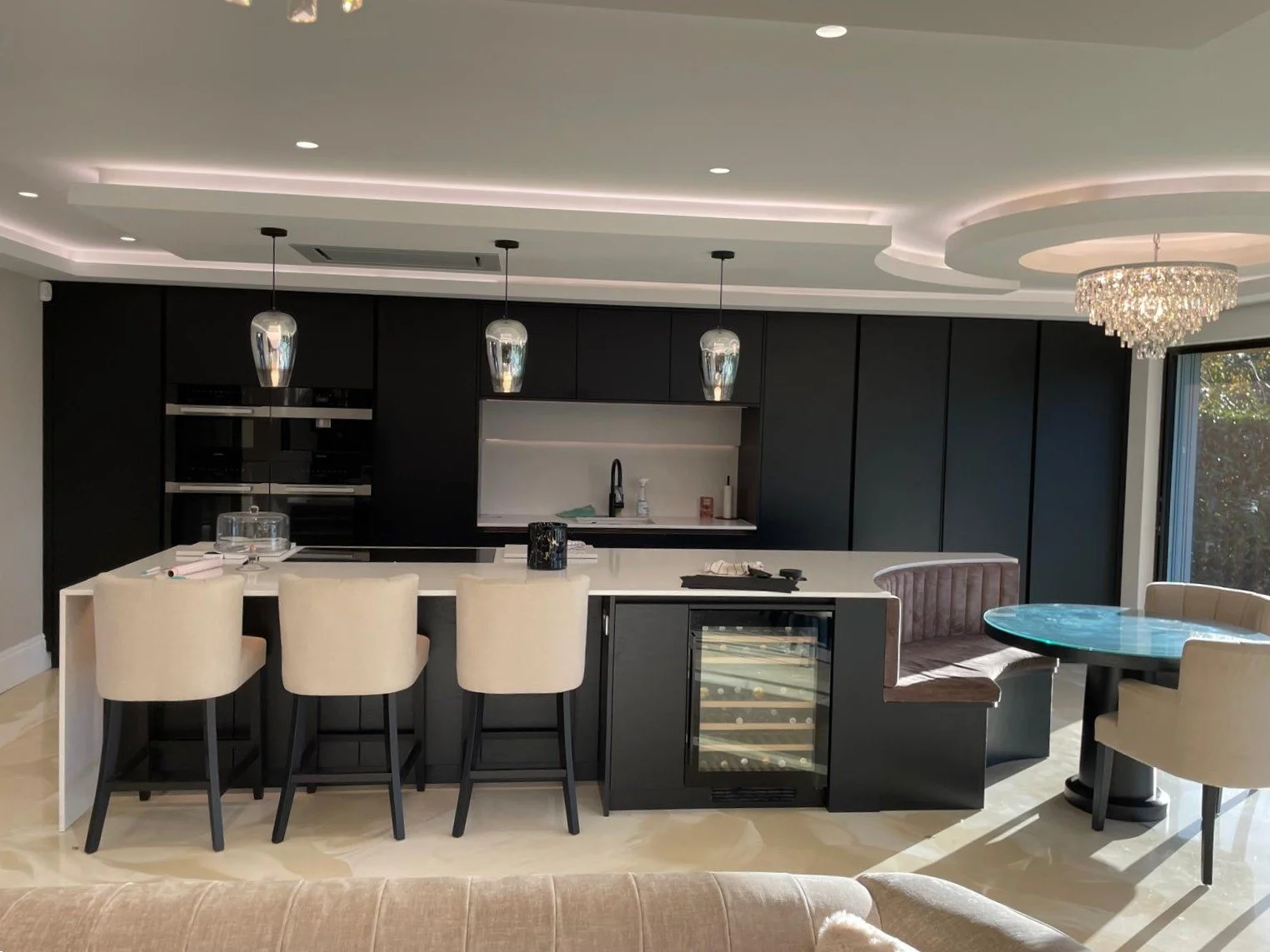 Modern kitchen with black cabinets, white island, beige chairs, and a glass table with a velvet booth and chandelier.