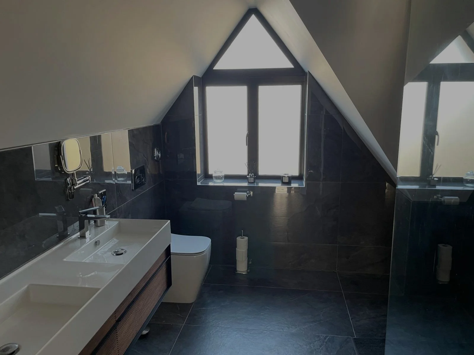 Modern bathroom with a sloped ceiling, black tiled walls and floor, a white sink with two faucets, a toilet, a toilet paper holder, and black window frames with a triangular and rectangular window letting in natural light.