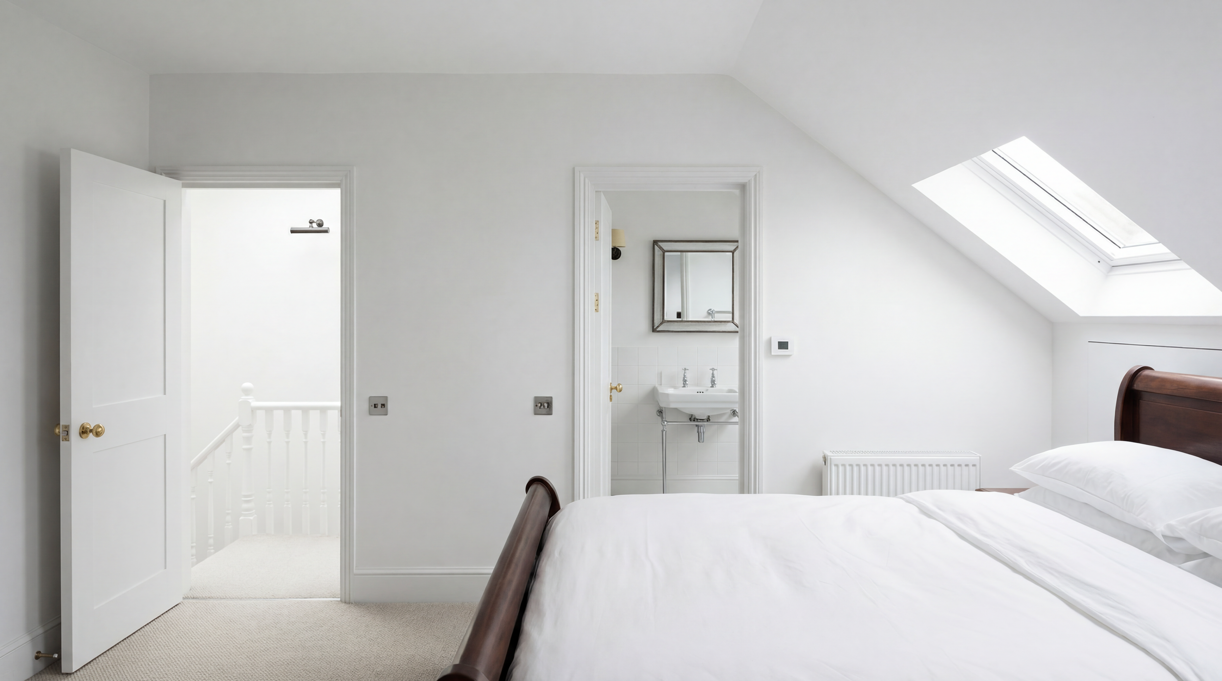 A bright bedroom with white walls, a skylight window, a wooden bed with white bedding, and an attached bathroom with a small sink and mirror.