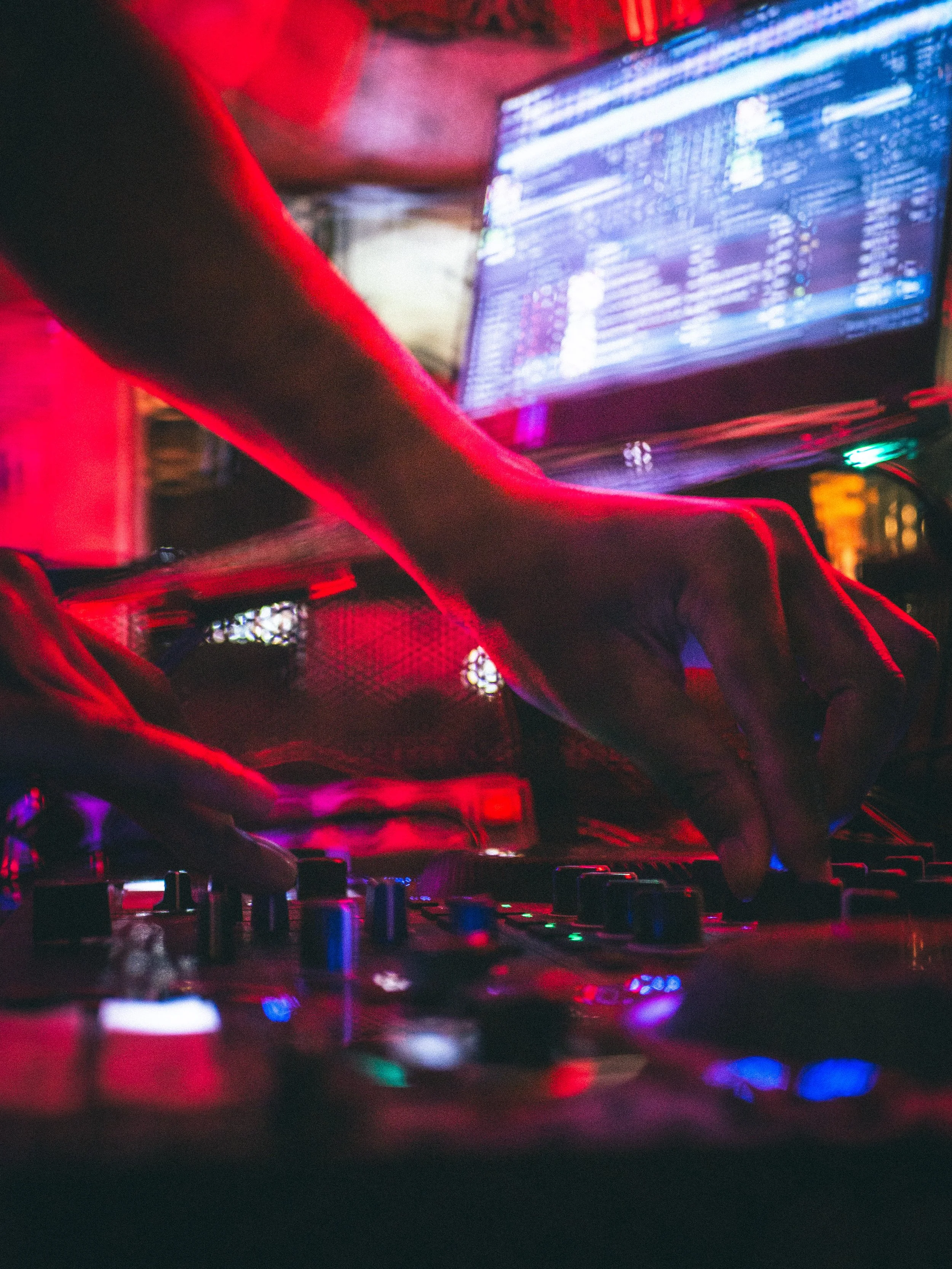 Main d'un DJ manipulant des contrôleurs de musique avec un écran d'ordinateur en arrière-plan, dans une ambiance de fête avec lumières rouges et bleues.
