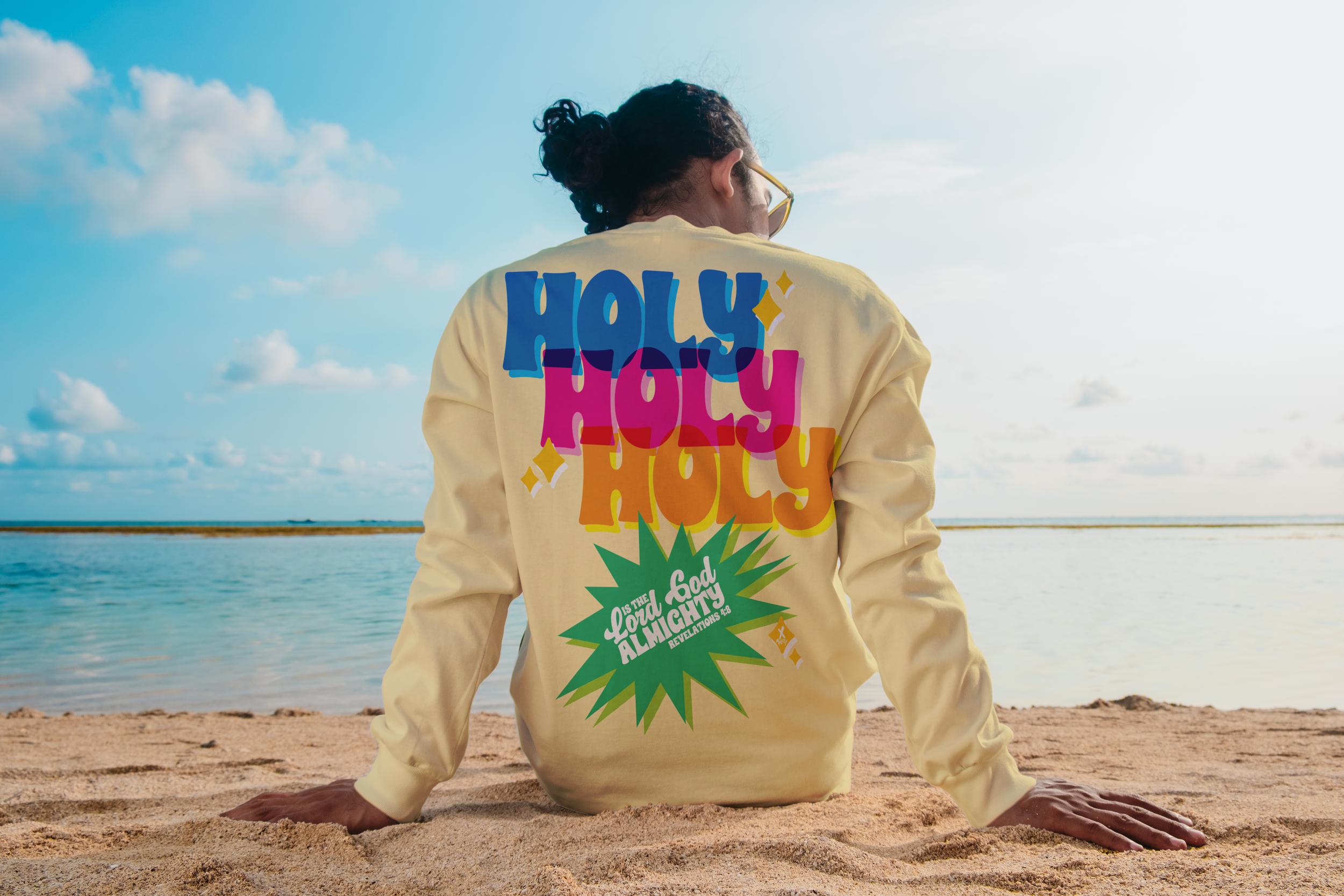Person sitting on sandy beach near water, wearing a yellow sweatshirt with colorful text and graphics on the back, facing away from the camera, with a partly cloudy sky.