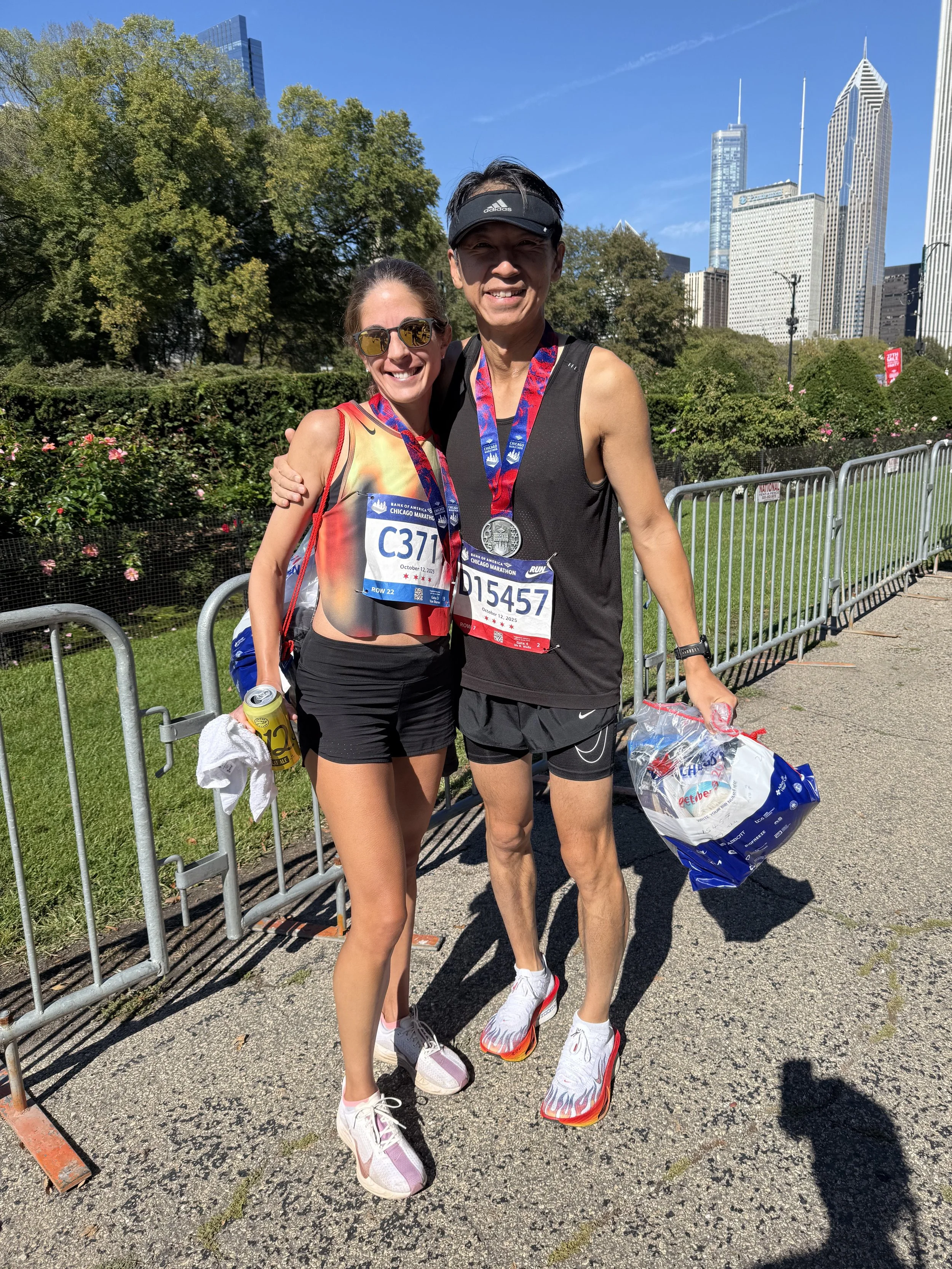 Two marathon runners, a woman and a man, smiling and posing together after finishing a race, wearing medals and athletic gear, with a city skyline and trees in the background.