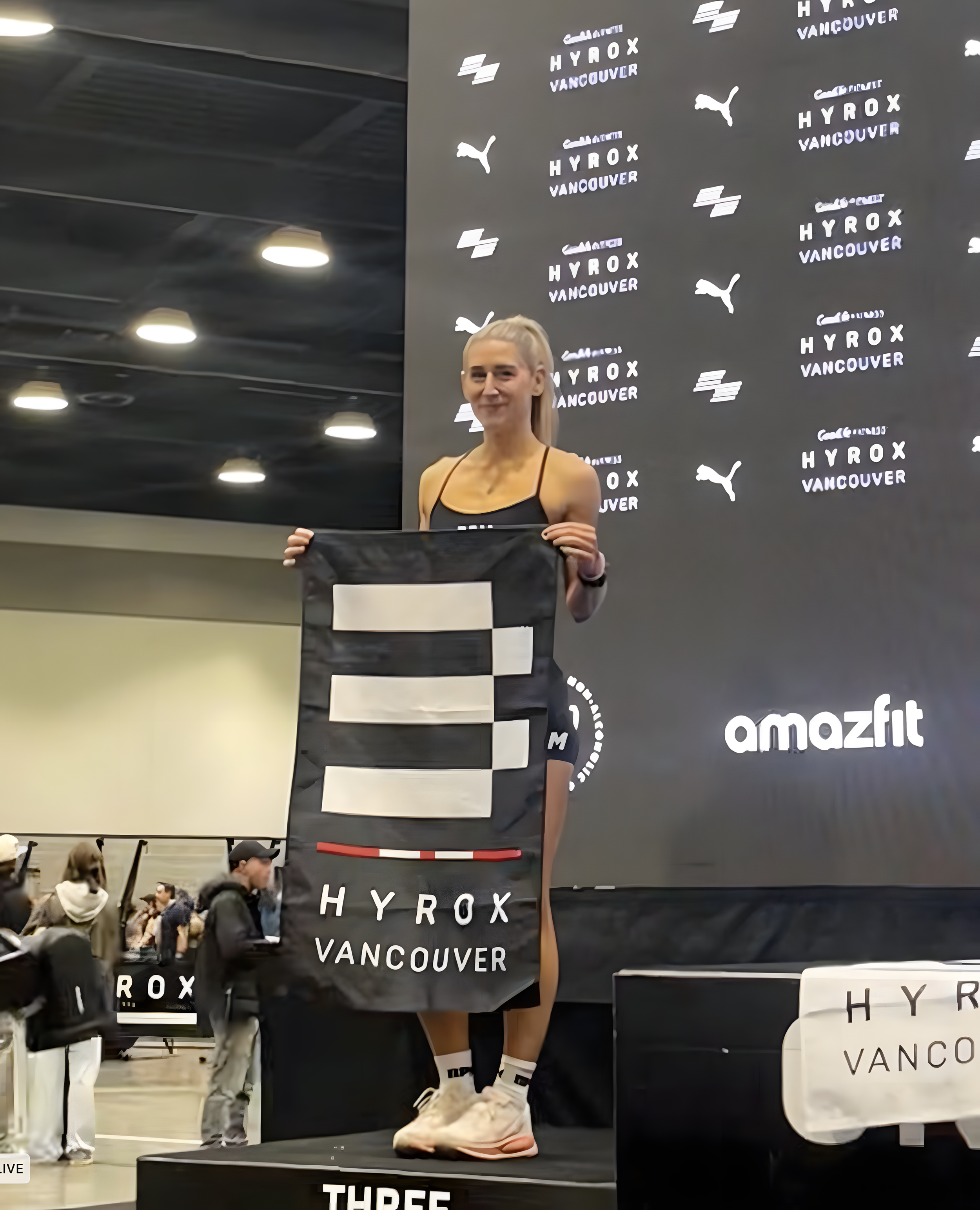 Woman holding a black and white prize banner at a Vancouver event, standing on a podium with a dark backdrop featuring logos of Hyrox Vancouver, Puma, and Amazfit.