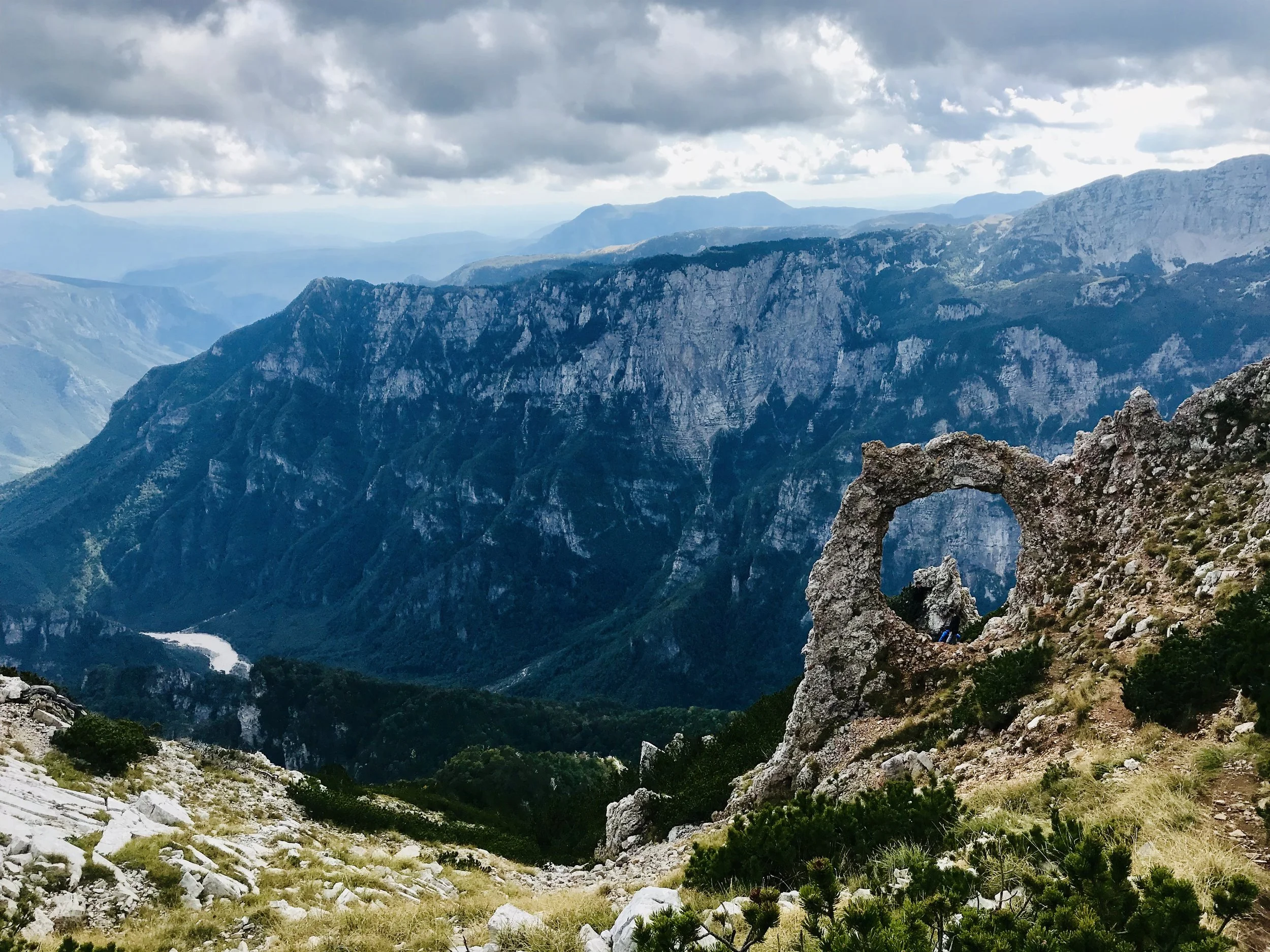 Mountain landscape with a natural stone arch in the foreground, surrounded by green shrubs and rocky terrain, under a cloudy sky.